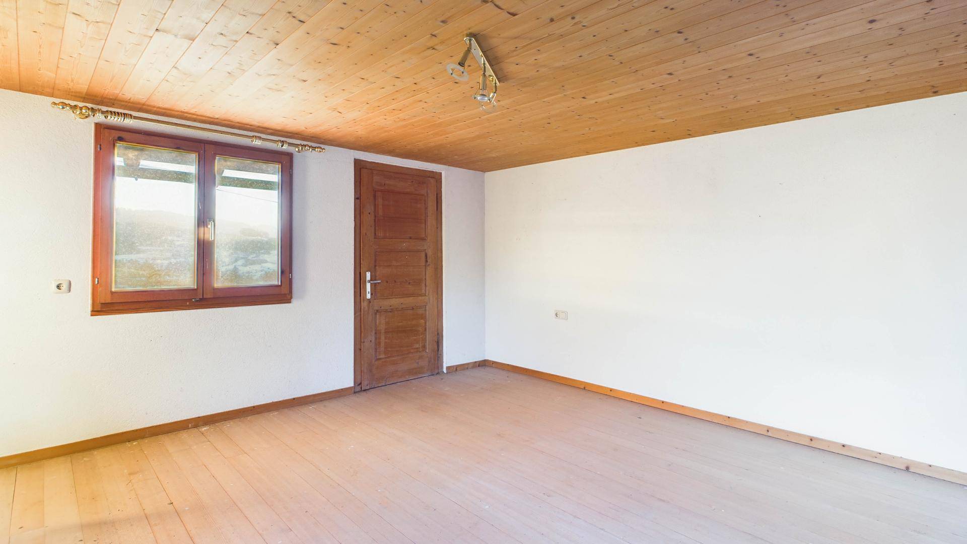 Leerer Raum mit Holzdielenboden und Holzdecke, durch ein Fenster fällt natürliches Licht herein.