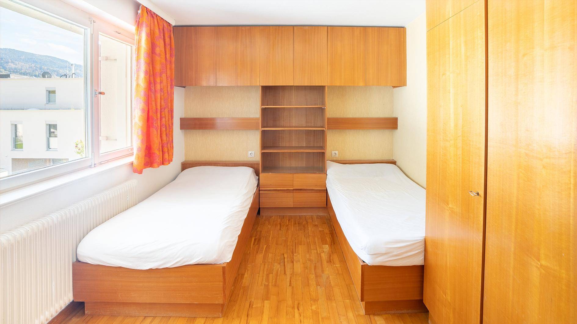 Einfaches Schlafzimmer mit zwei Betten, Holzschränken und einem Fenster.