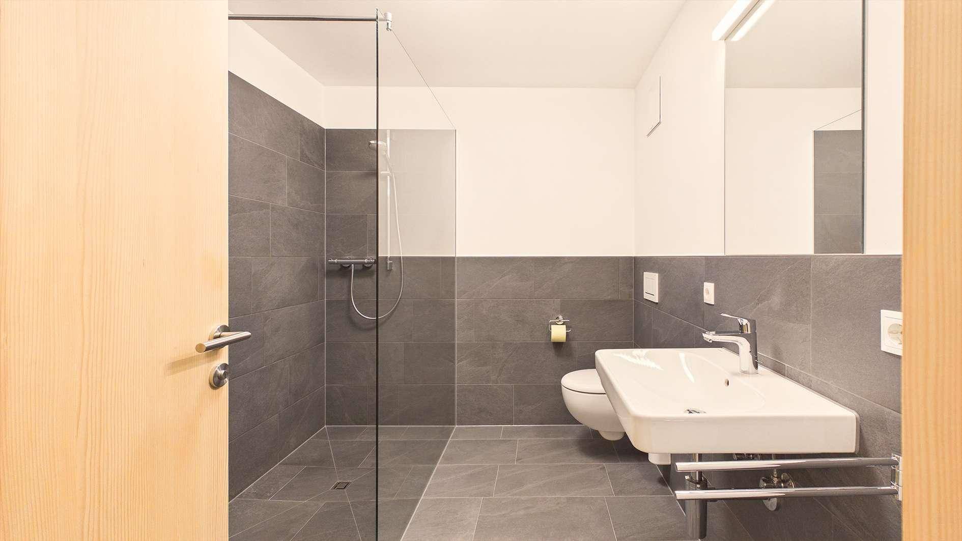 Modernes Badezimmer mit grauen Fliesen, ebenerdiger Dusche und wandhängendem WC.