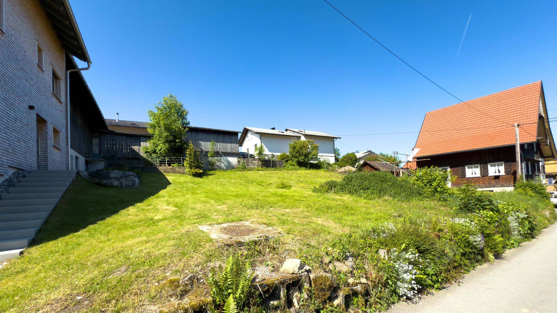 Blick auf ein grünes Grundstück neben einer Straße, umgeben von ländlichen Wohnhäusern.