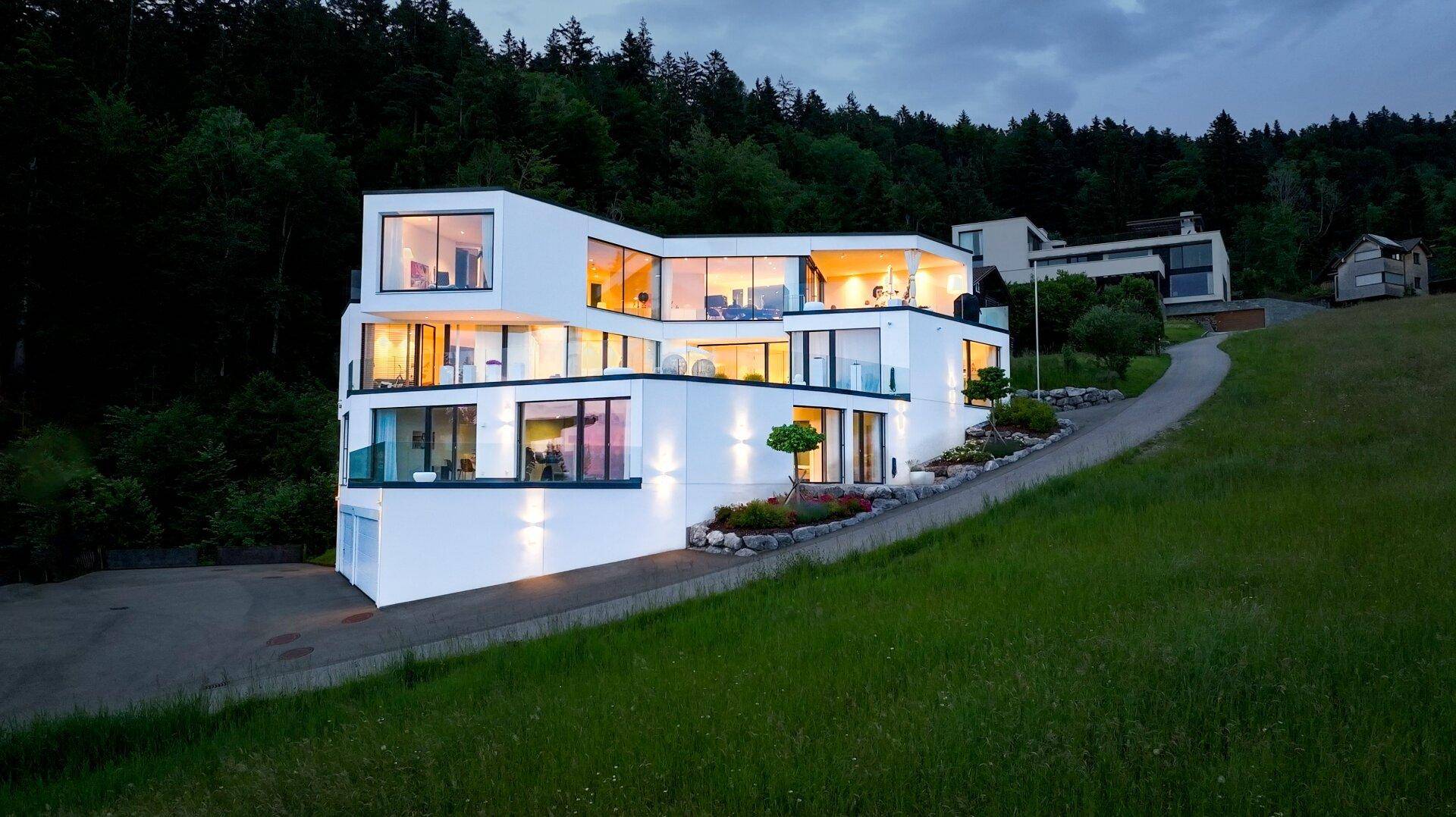 Moderne Villa mit markanter Architektur, großen Glasfronten und beleuchteten Terrassen in grüner Hanglage.