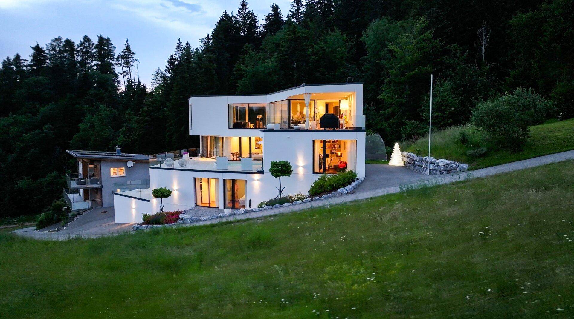 Moderne Villa mit markanter Architektur, großen Glasfronten und beleuchteten Terrassen in grüner Hanglage.