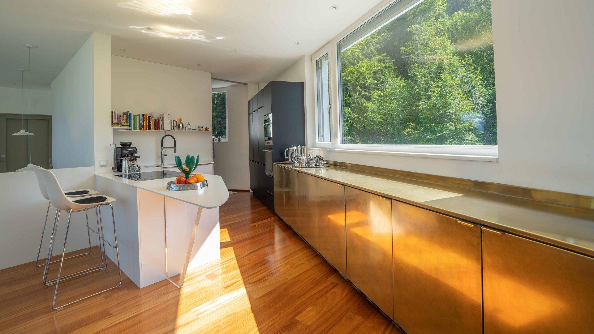 Moderne Küche mit goldenen Unterschränken, Kochinsel und großem Fenster mit Blick auf die Natur.