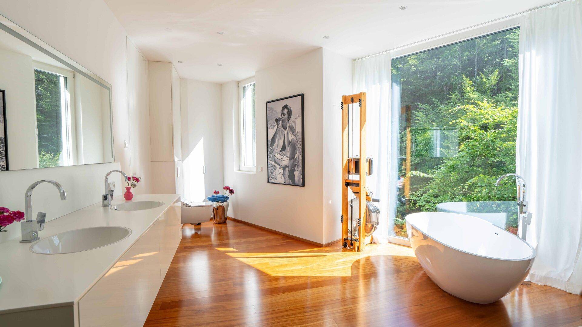 Helles Badezimmer mit freistehender Badewanne, Doppelwaschbecken und großem Fenster mit Blick ins Grüne.