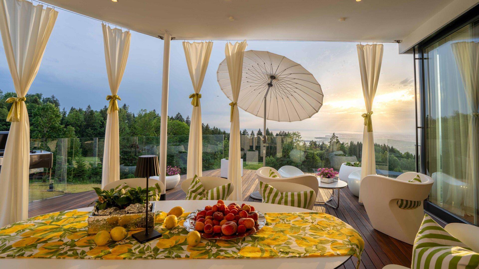 Überdachte Terrasse mit Esstisch, Vorhängen und weitem Blick über die grüne Landschaft und den See.