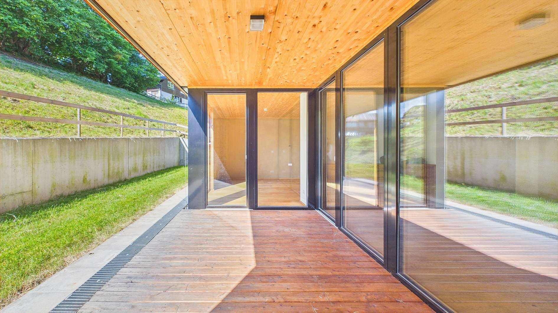 Geräumige Holzterrasse mit Glasfronten und Holzdecke, bietet einen hellen Außenbereich mit Blick ins Grüne.