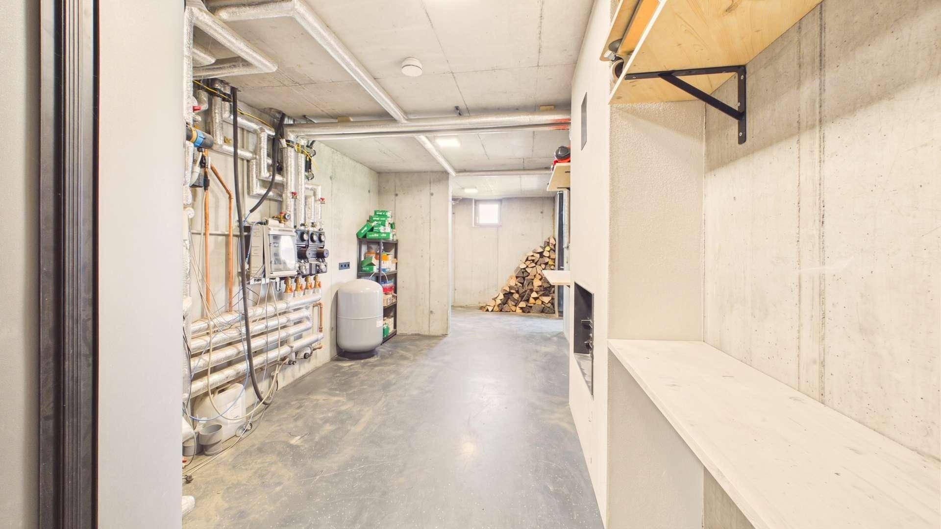Technikraum im Keller mit Heizungsanlagen, Rohren und einem Holzstapel für den Kamin.