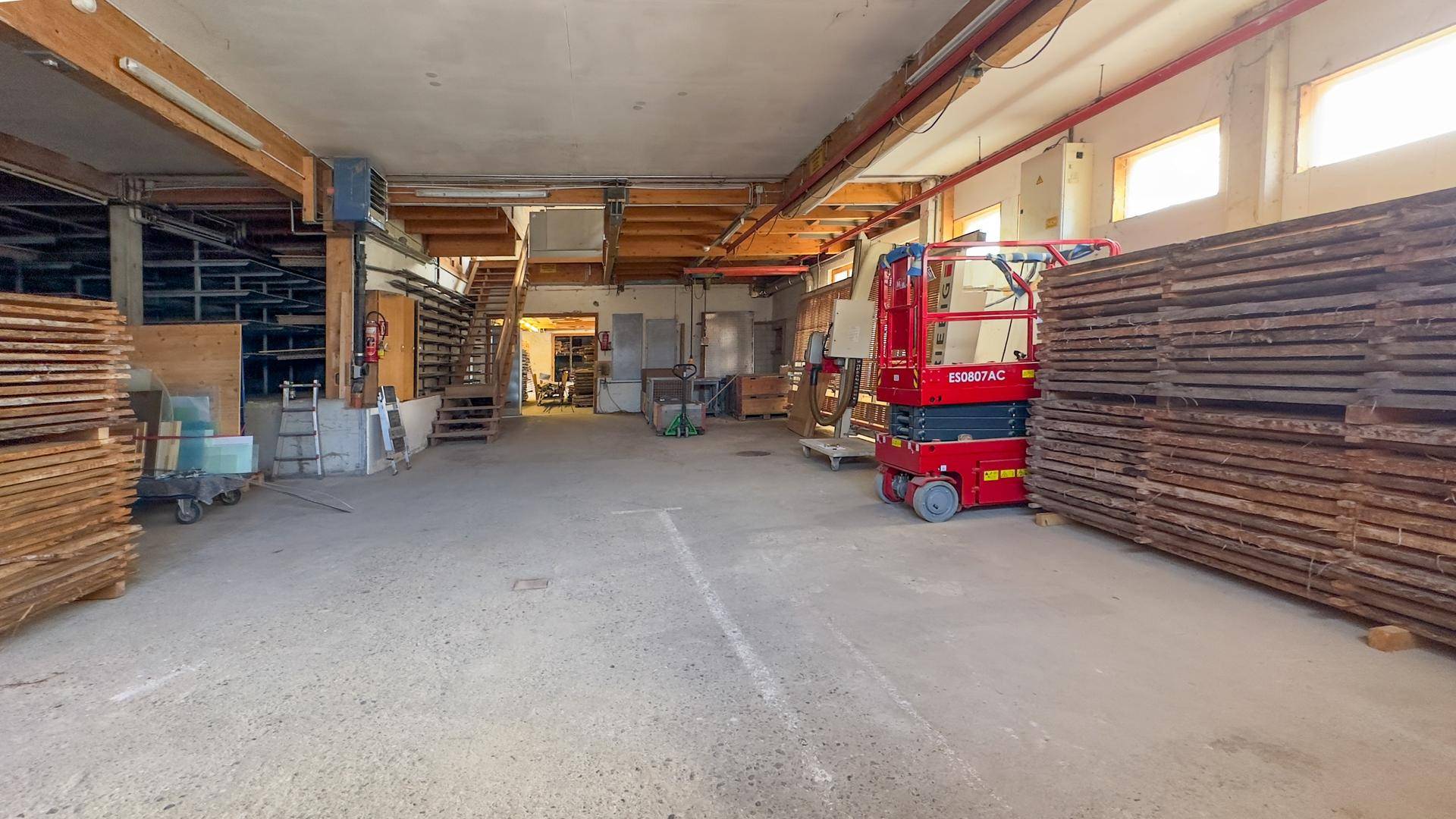 Weitläufiger Lagerraum mit Betonboden, hohen Decken und verschiedenen Geräten, darunter eine rote Hebebühne.