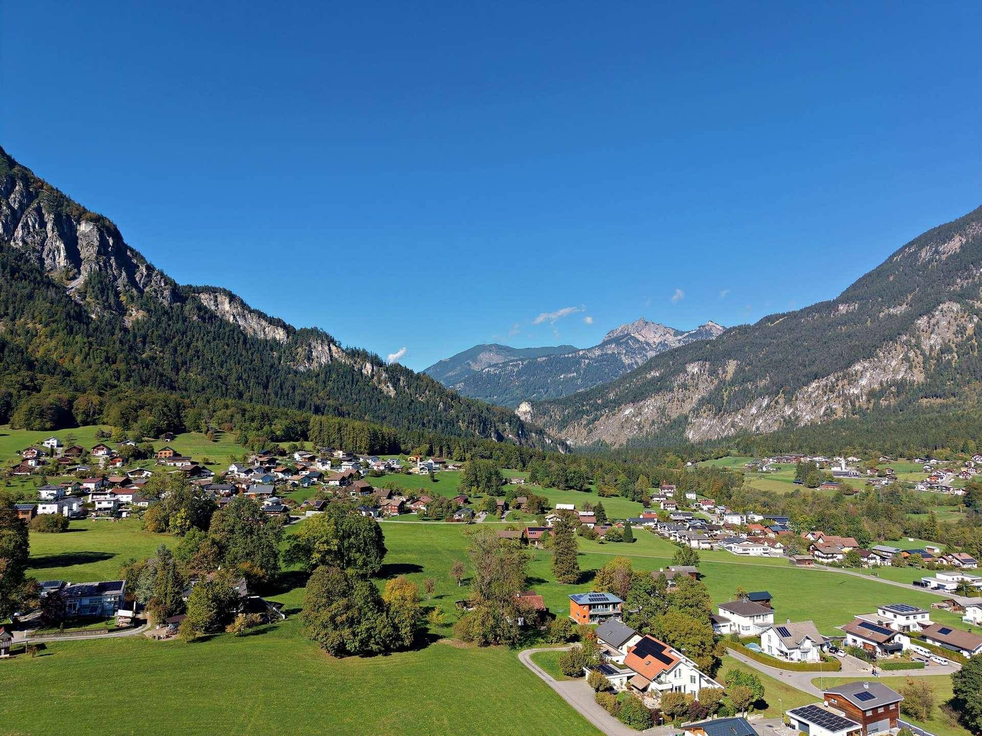 Panoramablick aus der Luft auf ein idyllisches Dorf umgeben von grünen Hügeln und Bergen.