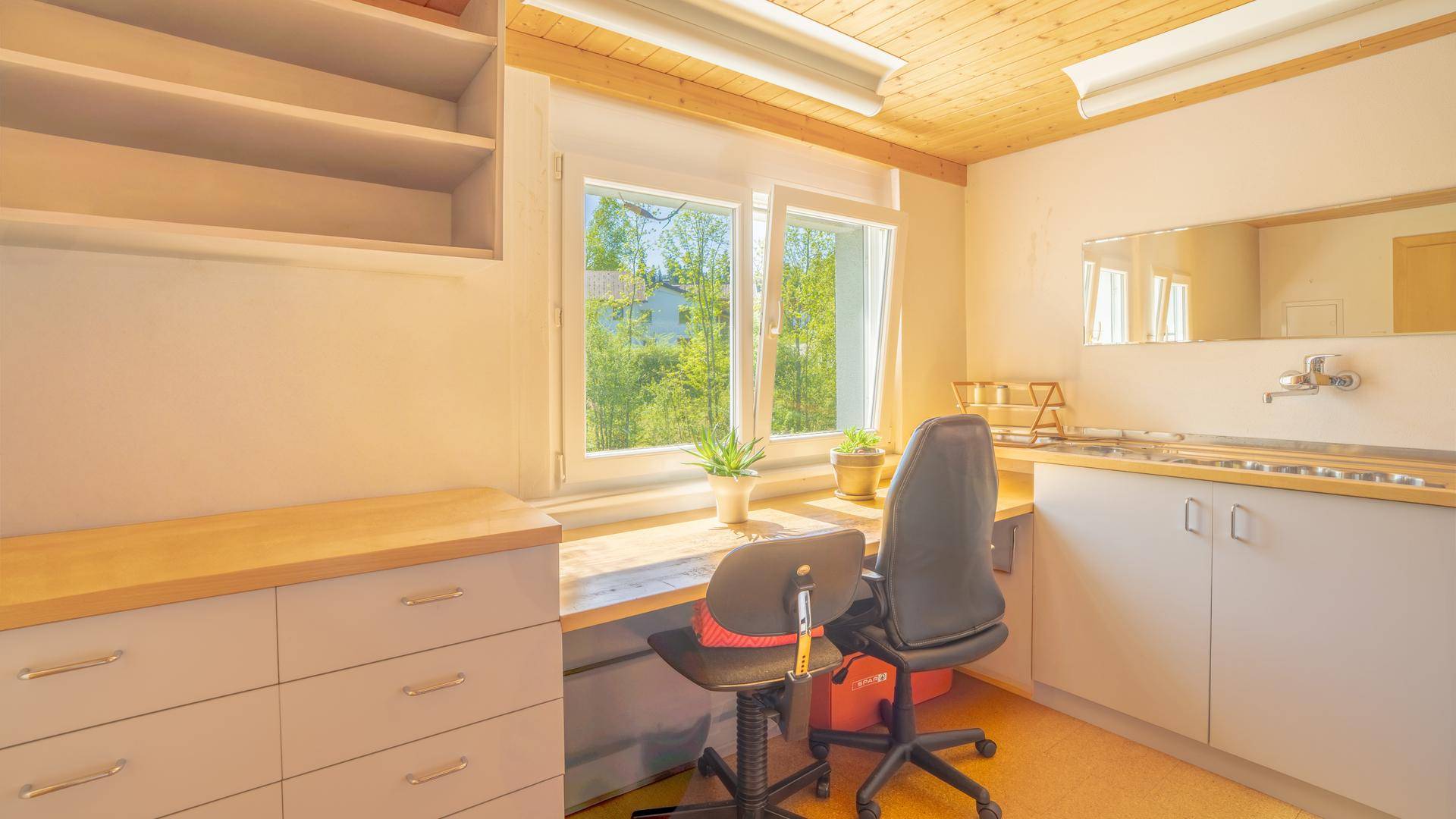 Helles Büro mit Arbeitsplatz, Regalen und kleiner Küchenzeile unter einer Holzdecke.