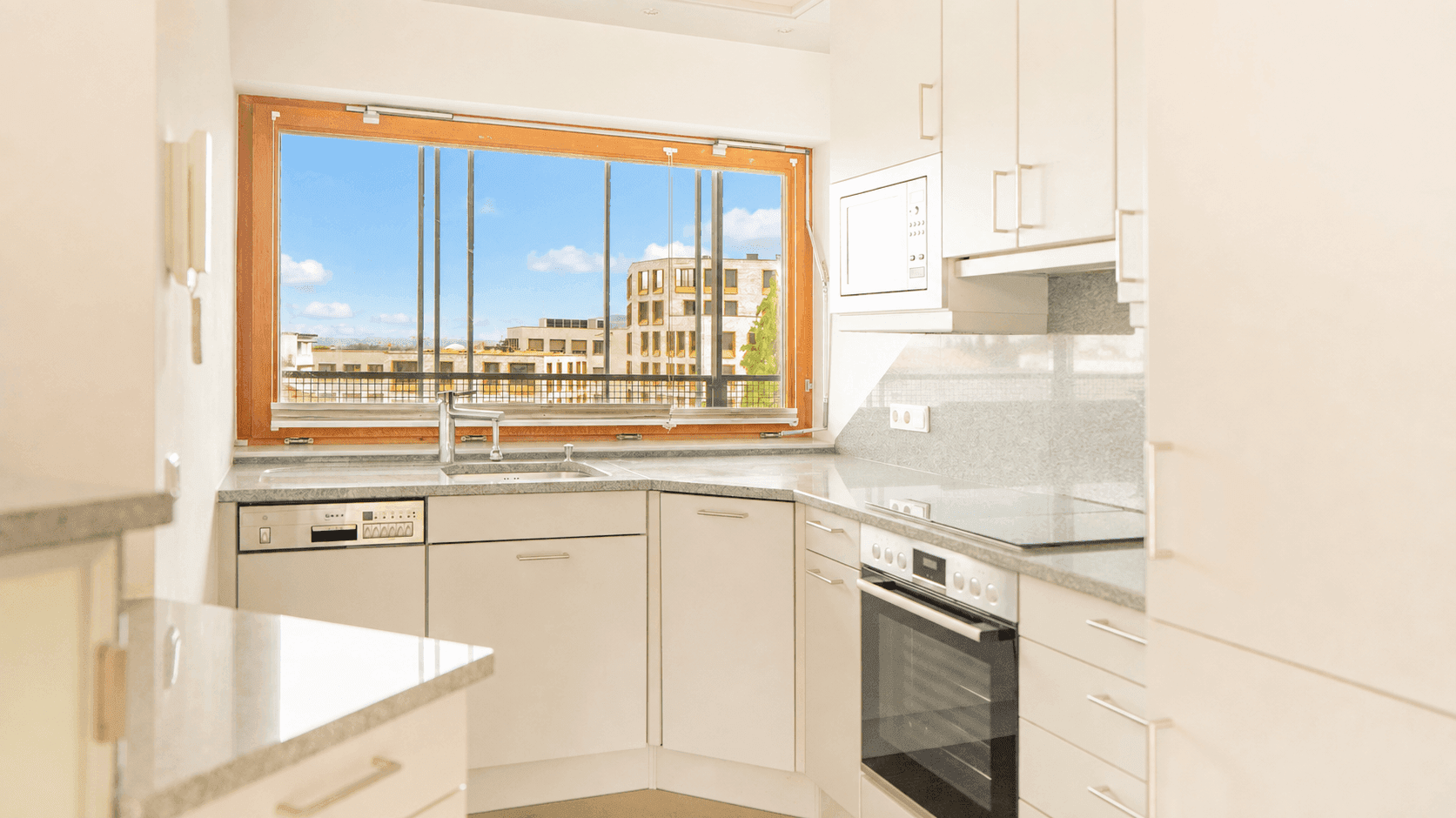 Moderne Einbauküche mit weißen Schränken, grauer Arbeitsplatte und Fenster mit Stadtblick.
