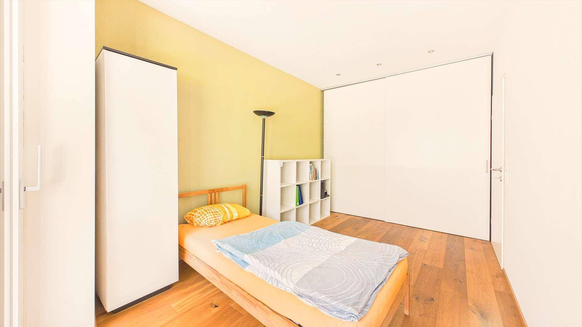Helles Schlafzimmer mit Holzboden, gelber Akzentwand und Einzelbett.