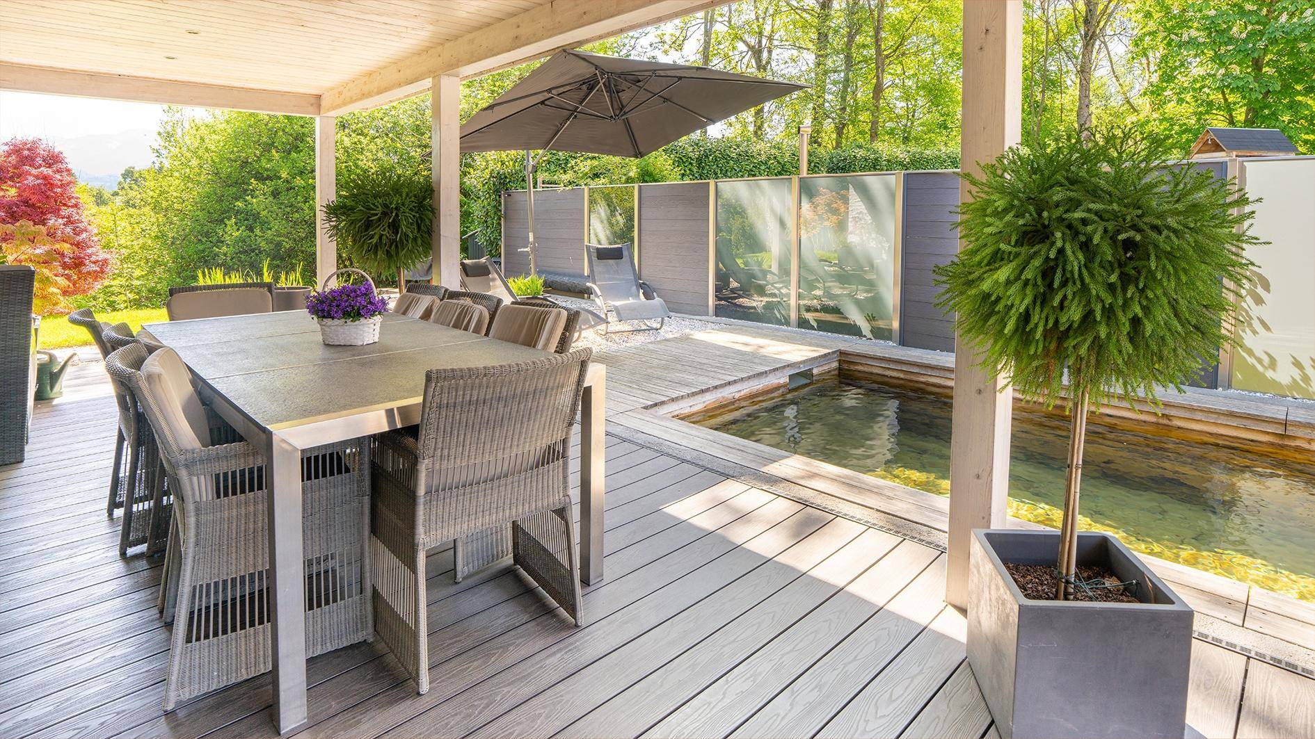 Große überdachte Terrasse mit Esstisch und Pflanzen, umgeben von viel Grün.
