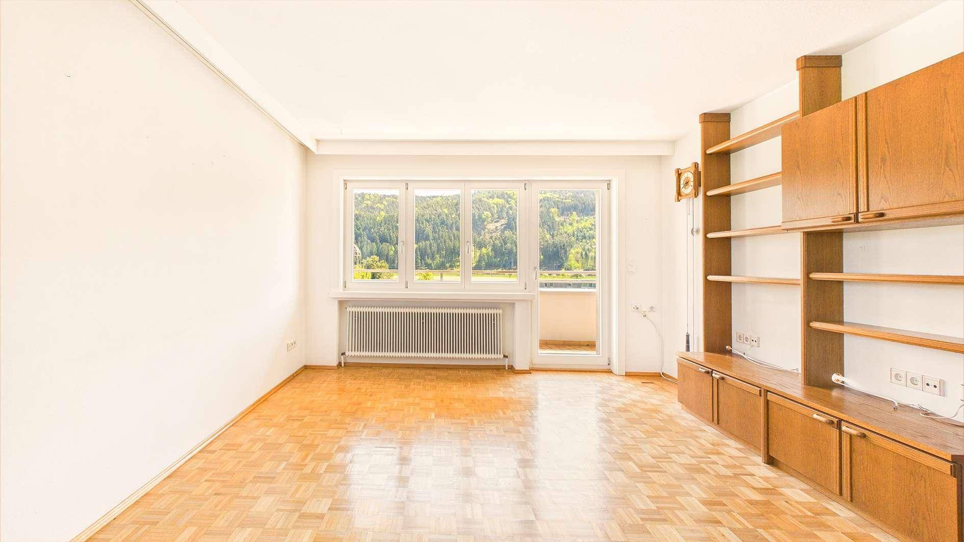 Geräumiges Wohnzimmer mit Parkettboden, Einbauschränken und Zugang zum Balkon mit Waldblick.