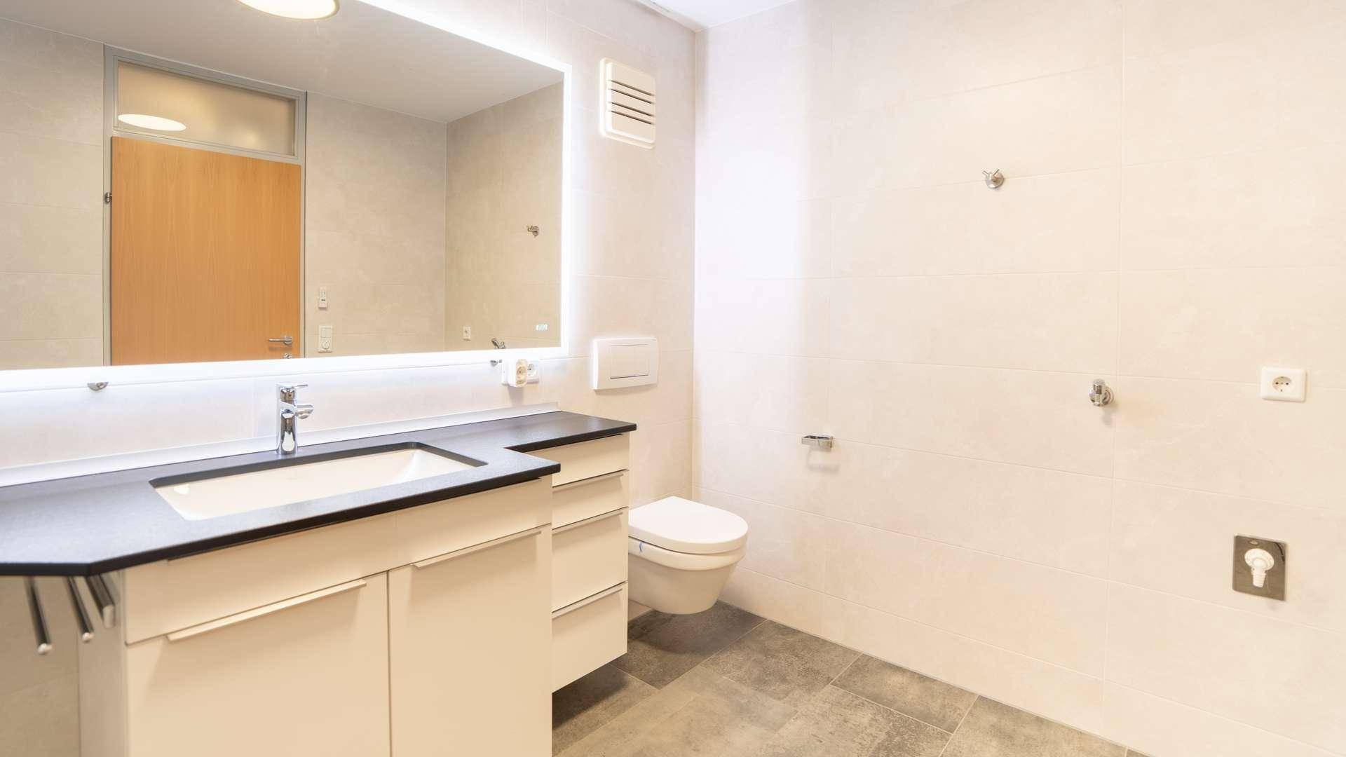 Helles Badezimmer mit WC, modernem Waschtisch, großem beleuchtetem Spiegel und Abluftventilator.