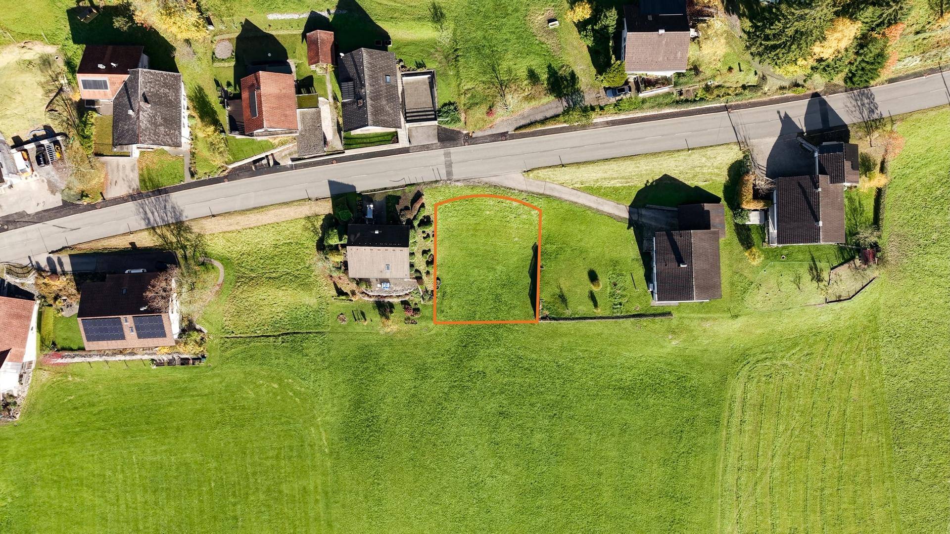 Luftaufnahme eines Baugrundstücks, umrandet in Orange, umgeben von Wohnhäusern und einer Straße.