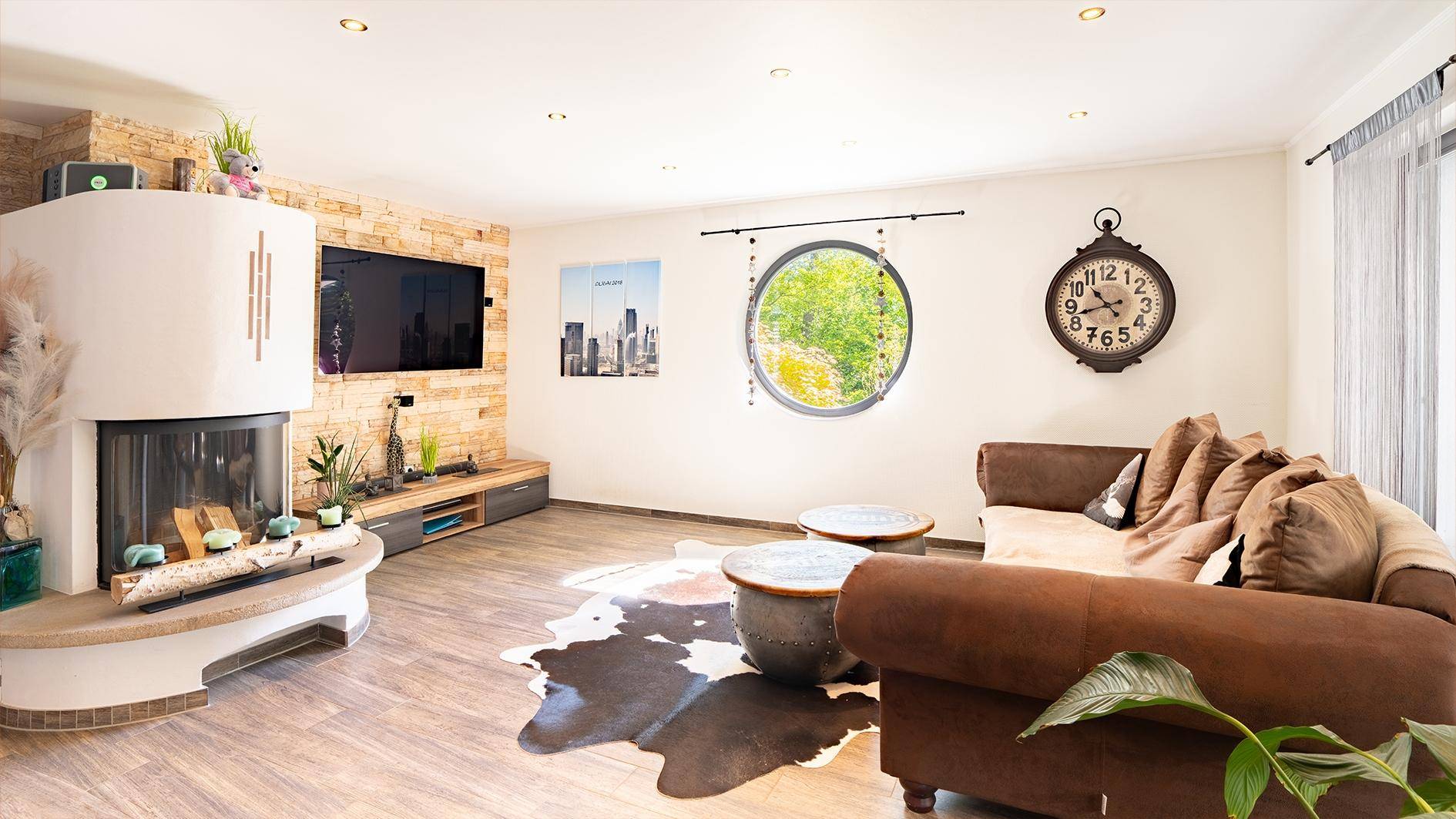 Gemütliches Wohnzimmer mit Kamin, Holzboden und einem runden Fenster mit Blick ins Grüne.