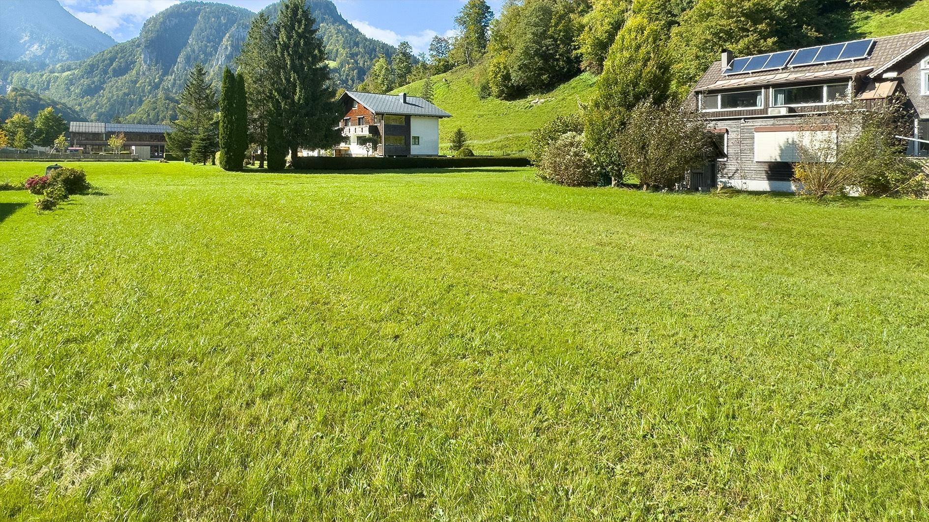 Weitläufige grüne Wiese mit Blick auf umliegende Häuser und die beeindruckende Berglandschaft.