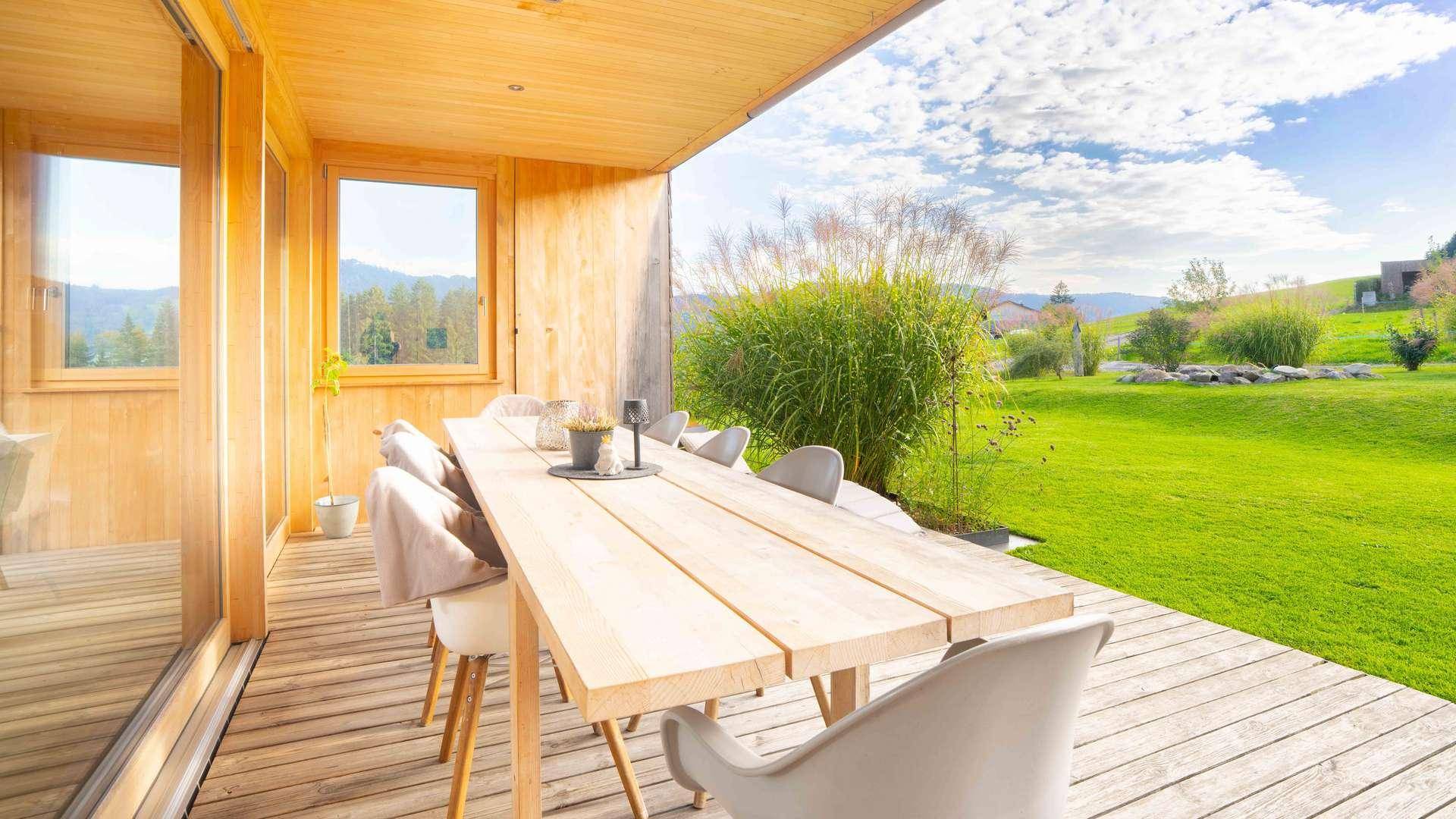 Überdachte Holzterrasse mit großem Esstisch und Stühlen, umgeben von grüner Landschaft.