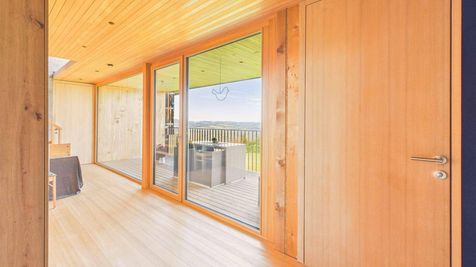 Heller Innenraum mit Holzwänden und großen Glasschiebetüren, die zu einem Balkon mit Aussicht führen.