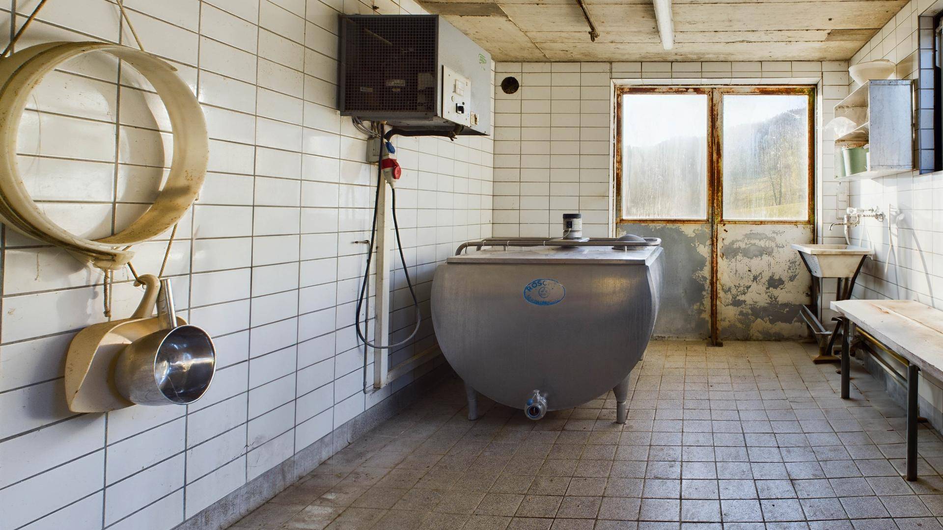 Gefliester Wirtschaftsraum mit großem Metalltank und Fenster, möglicherweise eine ehemalige Milchkammer.