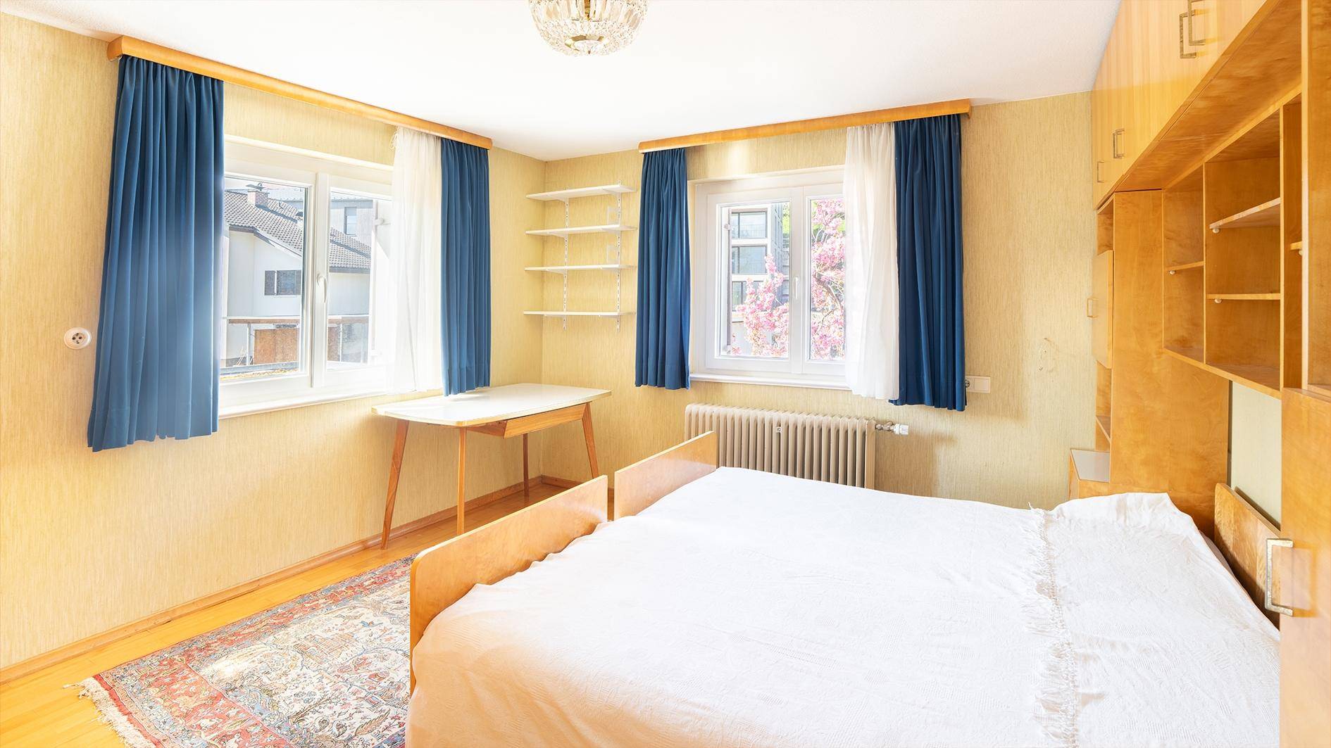 Schlafzimmer mit zwei Fenstern, blauen Vorhängen und integrierten Holzschränken.