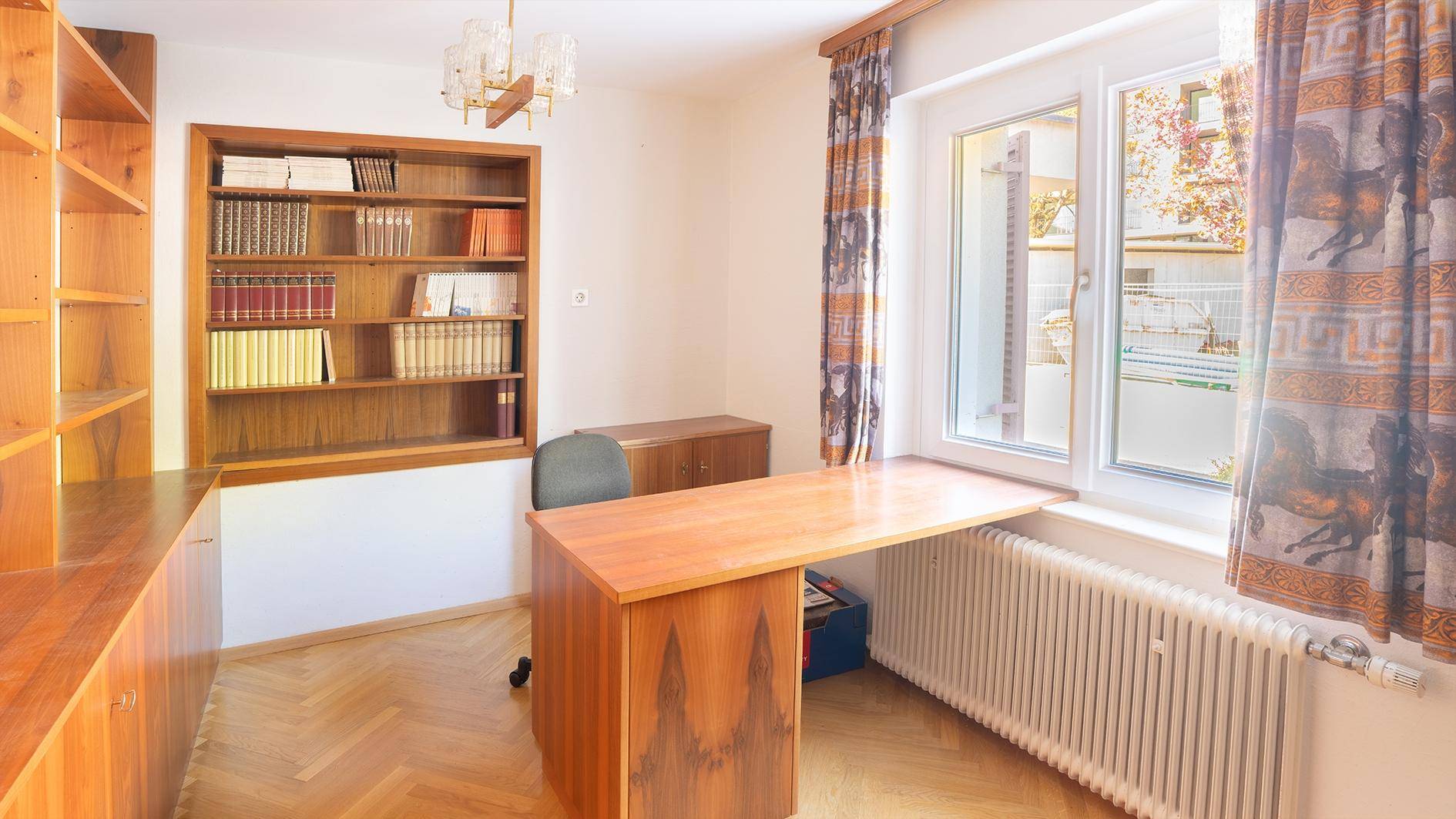 Helles Arbeitszimmer mit Schreibtisch, Bücherregal und Fenster mit Blick nach draußen.