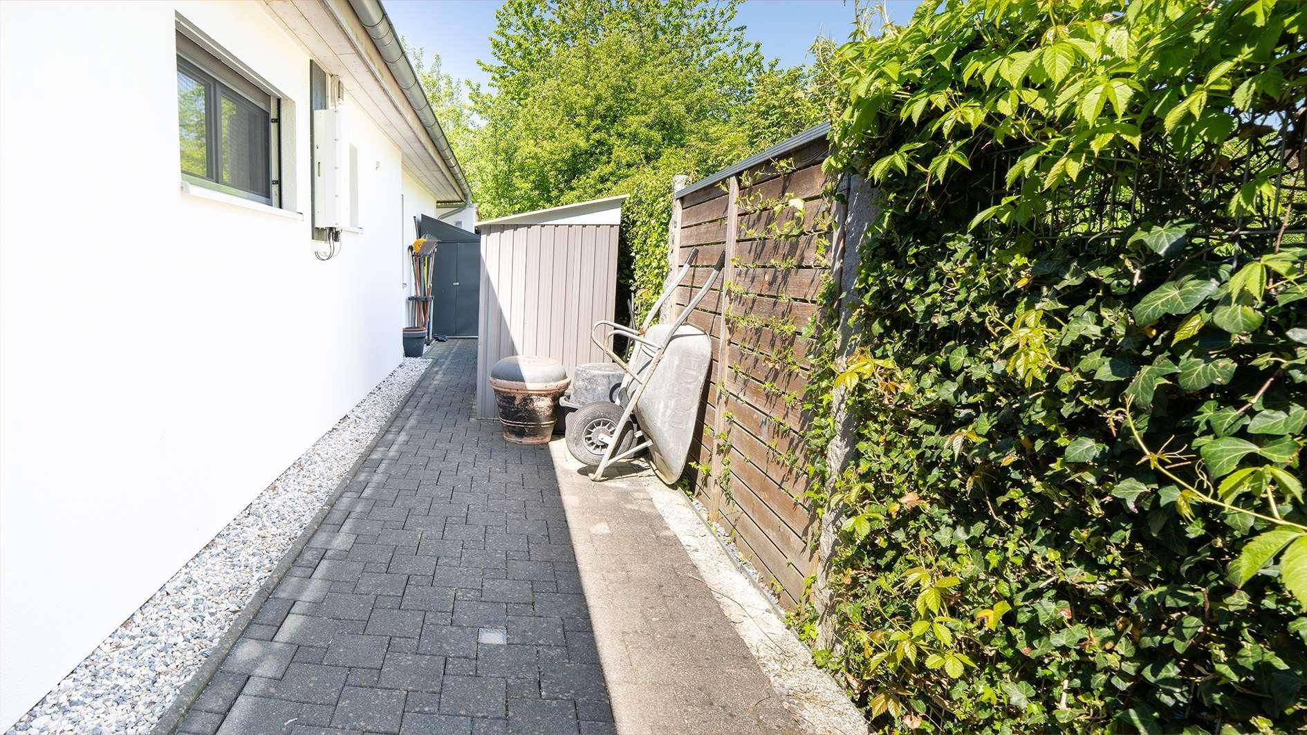 Schmaler gepflasterter Weg entlang der Hausseite mit Gartenschuppen und bewachsenem Zaun.