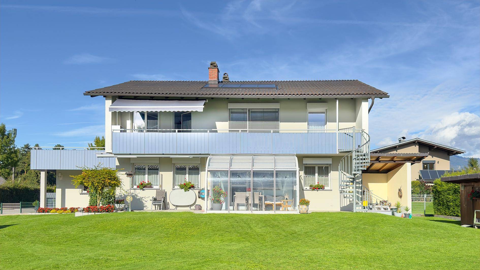 Freistehendes Haus mit großem Rasen, Balkonen, Wintergarten und Solaranlage unter blauem Himmel.