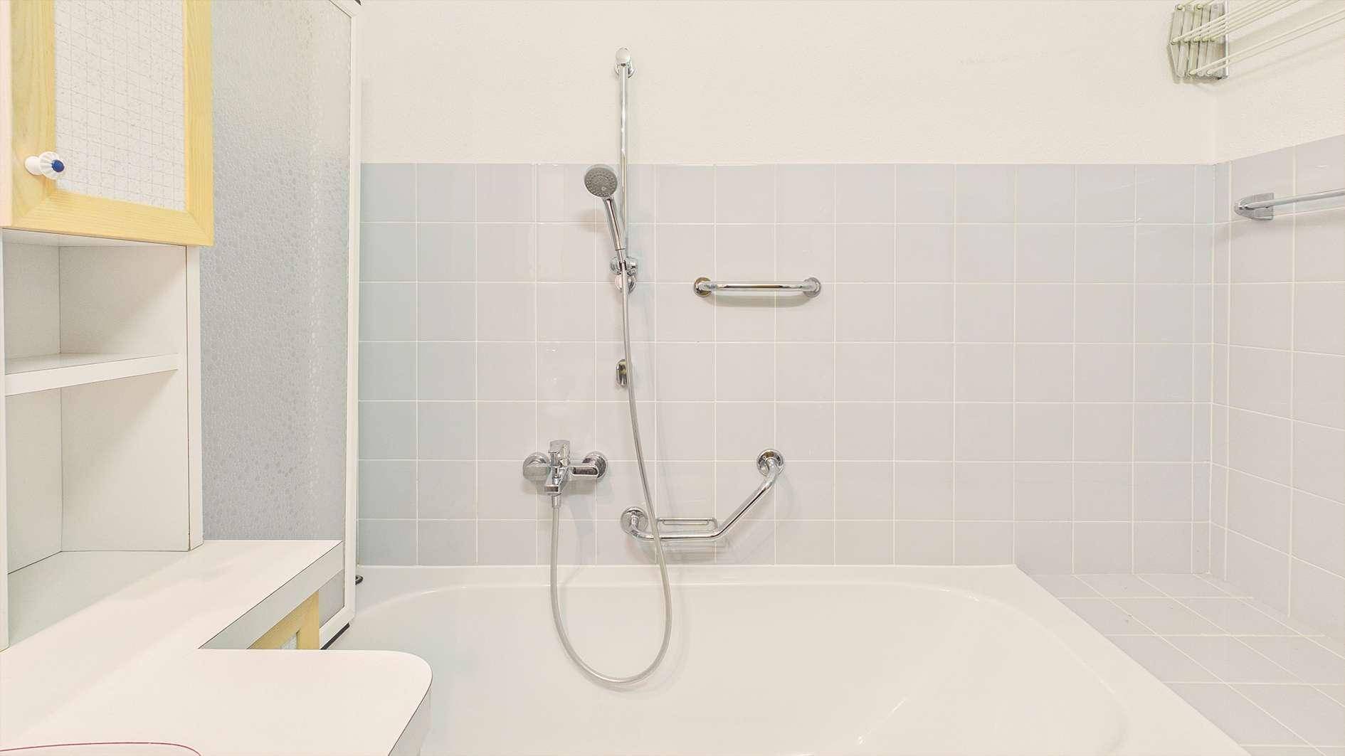 Badewanne mit Duscharmatur und Haltegriffen in einem hell gefliesten Badezimmer.
