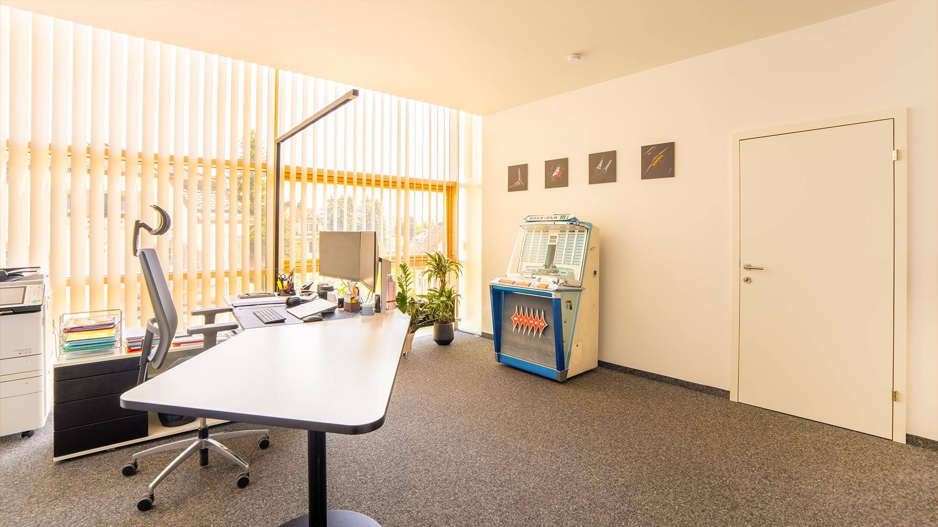 Modernes Büro mit Schreibtisch, Bürostuhl und einem dekorativen Musikautomaten, viel Tageslicht durch Jalousien.
