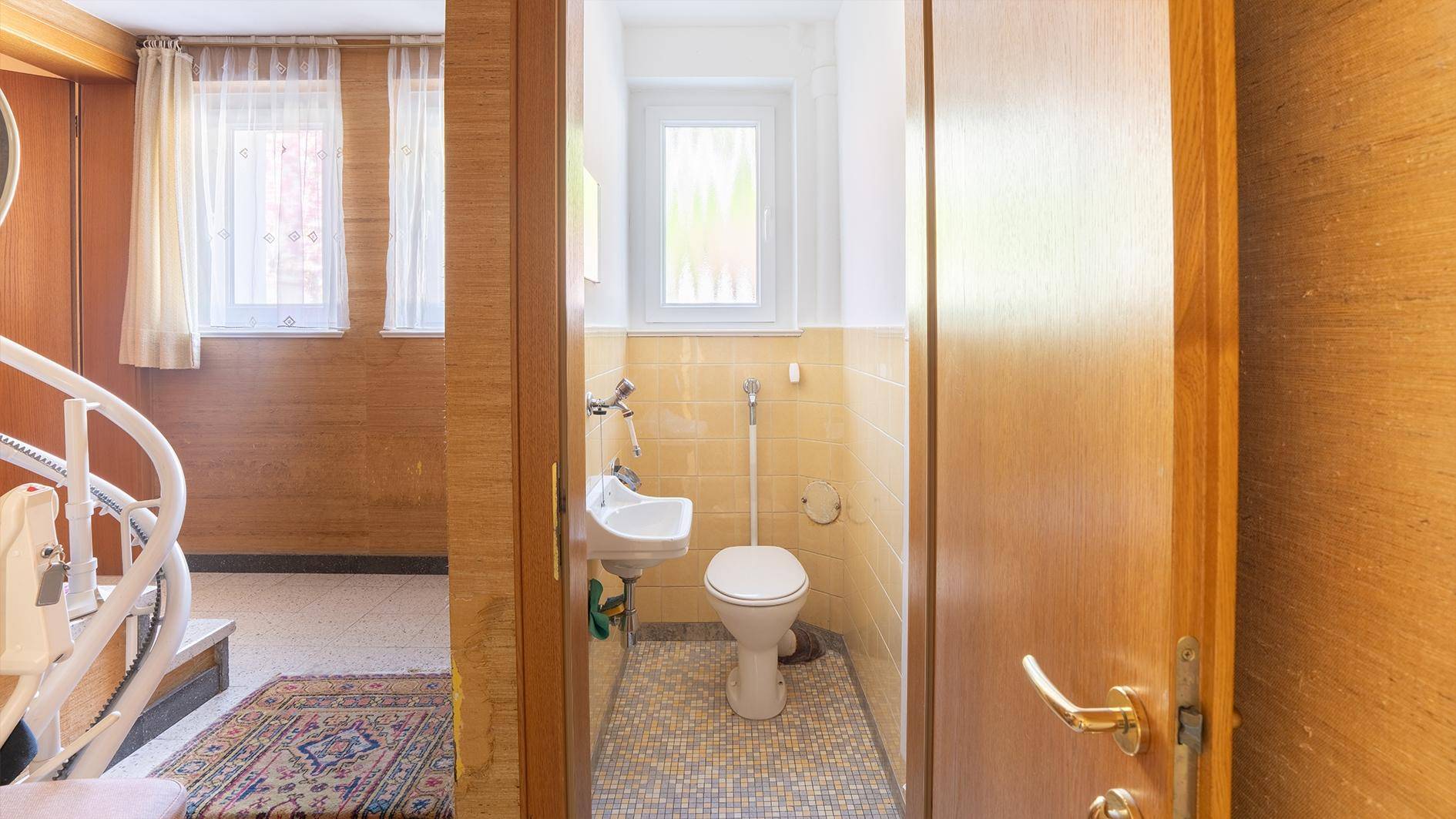 Kleines Gäste-WC mit gelben Fliesen und Waschbecken, vom Flur mit Treppenlift aus sichtbar.
