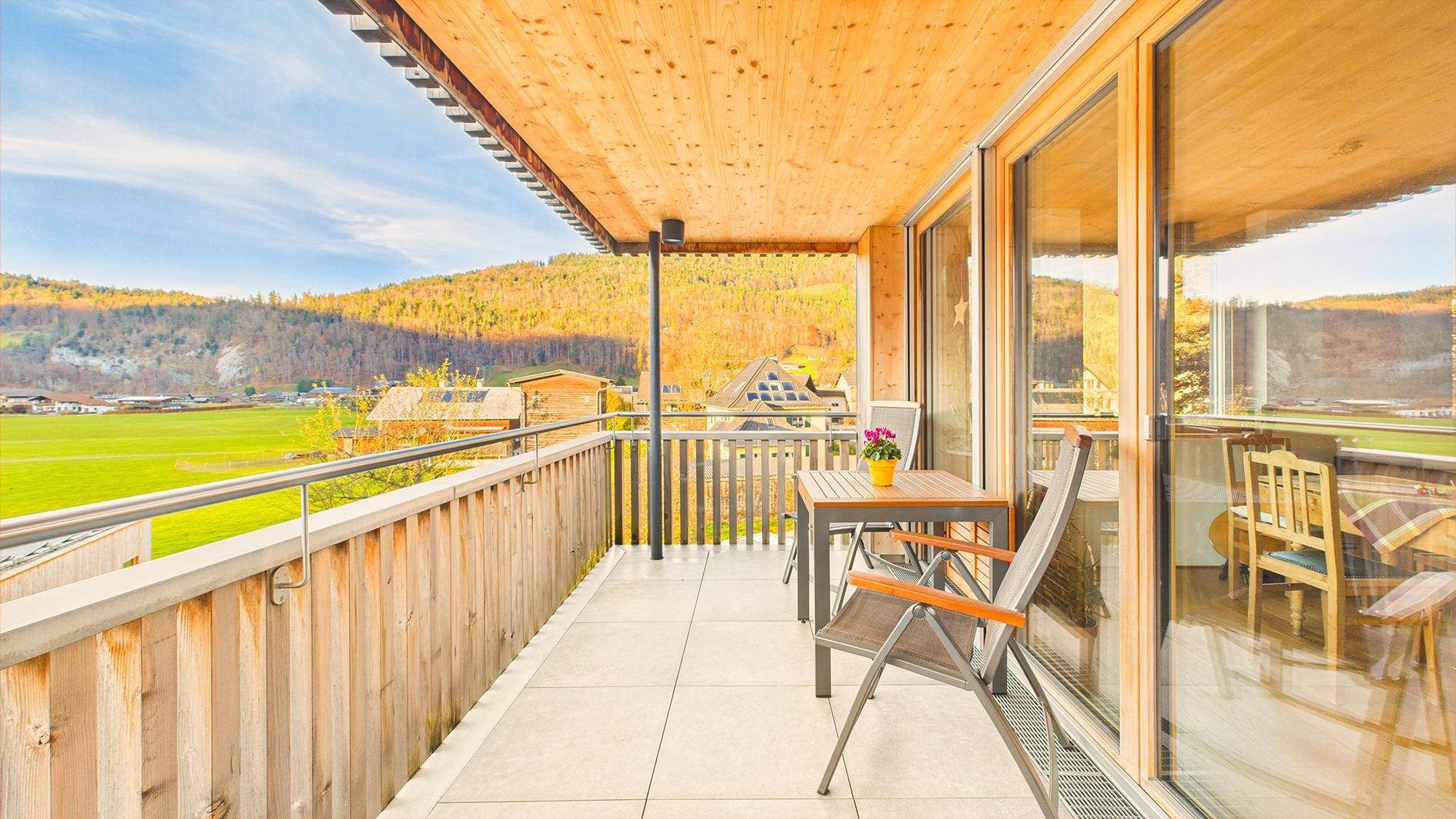 Sonniger Balkon mit Holzboden, Sitzgelegenheiten und weitem Blick auf die grüne Landschaft.
