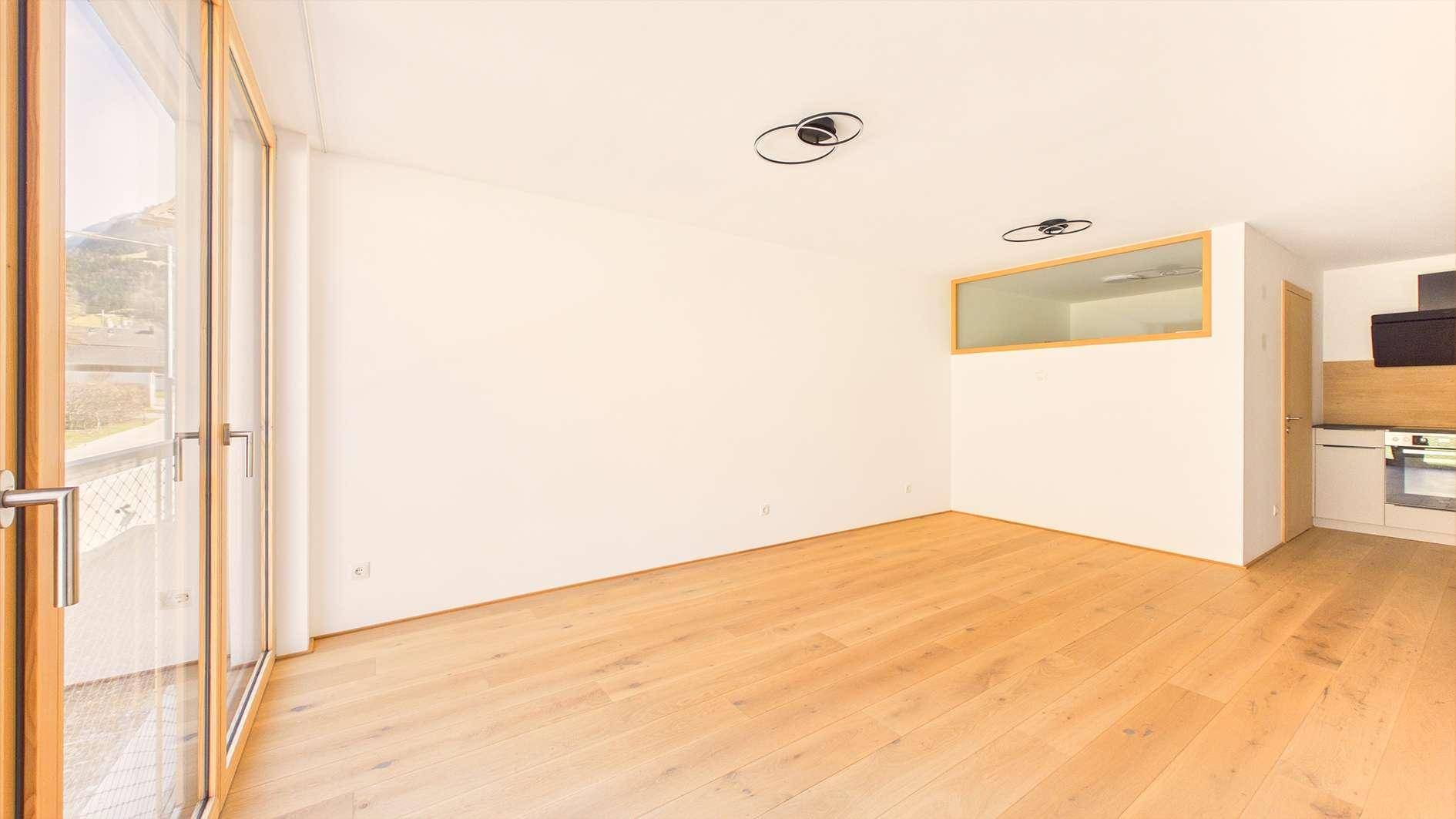 Geräumiger, leerer Wohnbereich mit Holzboden und großen Fenstern, die viel Licht hereinlassen.