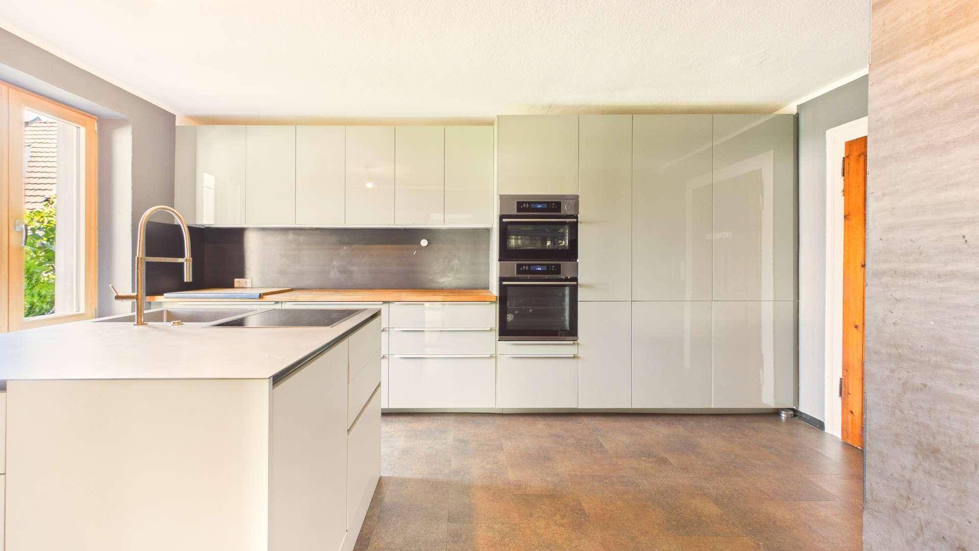 Moderne Küche mit glänzenden hellgrauen Fronten, Kochinsel und integrierten Backöfen.