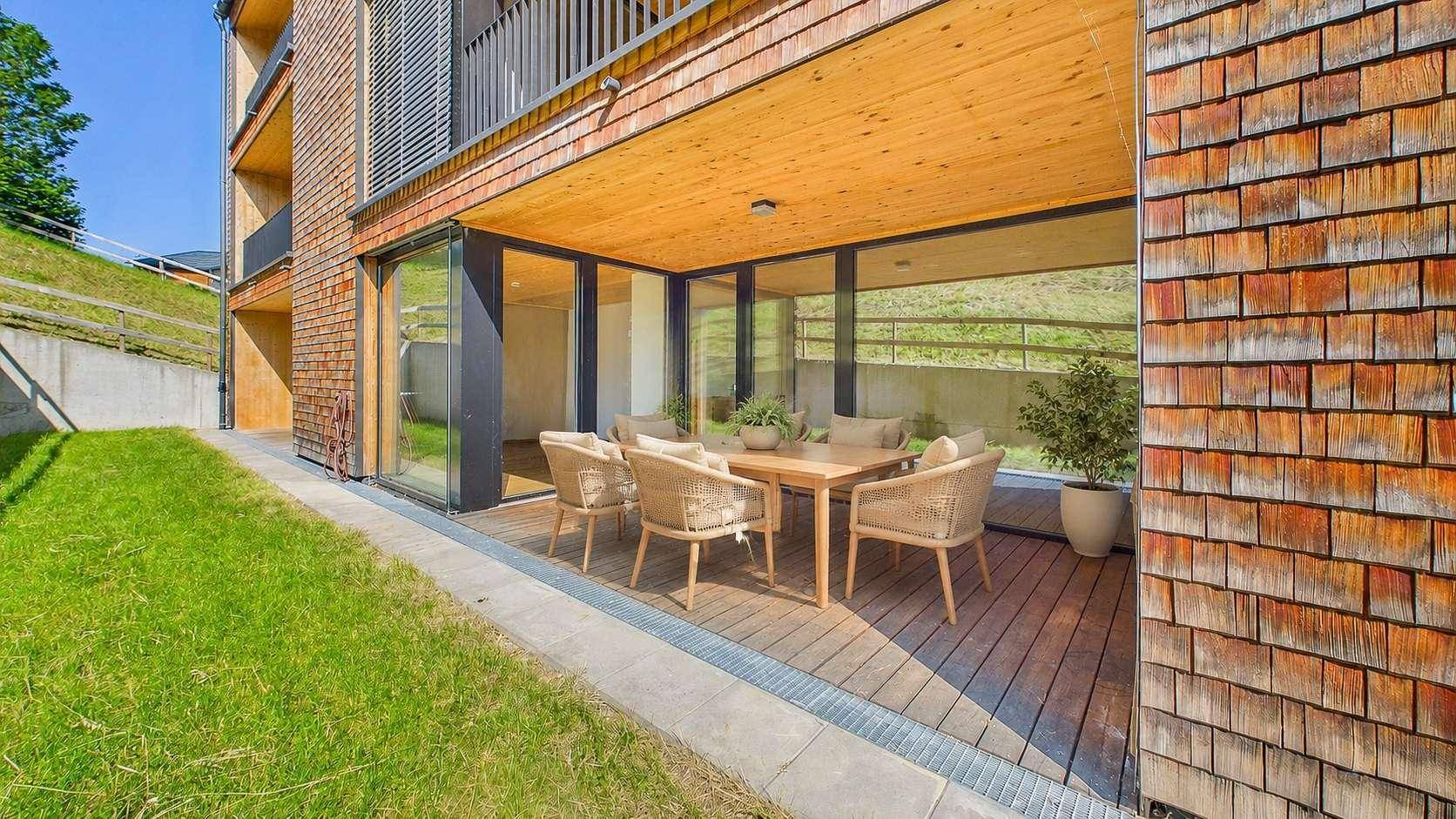 Möblierte Holzterrasse mit Sitzgelegenheiten und Blick ins Grüne, angrenzend an ein Gebäude mit Holzschindelfassade.