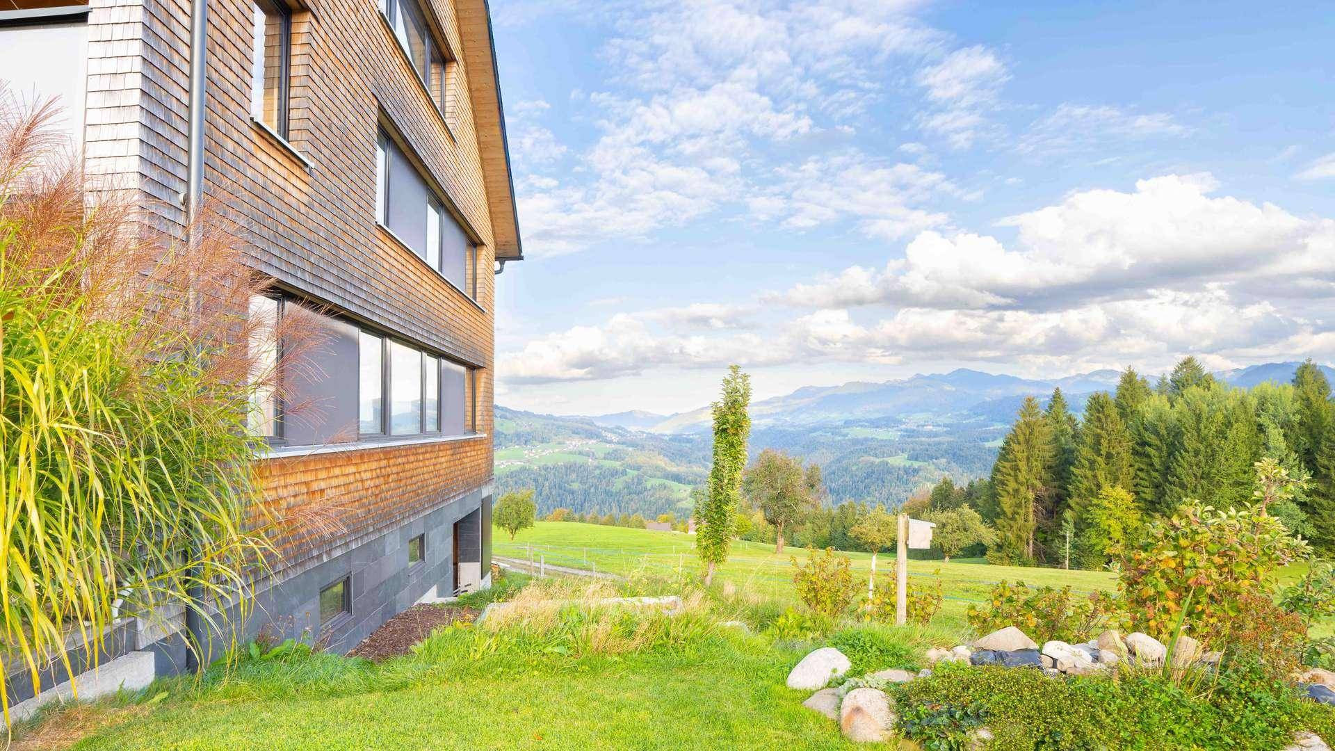 Seitenansicht des Holzhauses mit großen Fenstern und Panoramablick auf die grüne Berglandschaft.