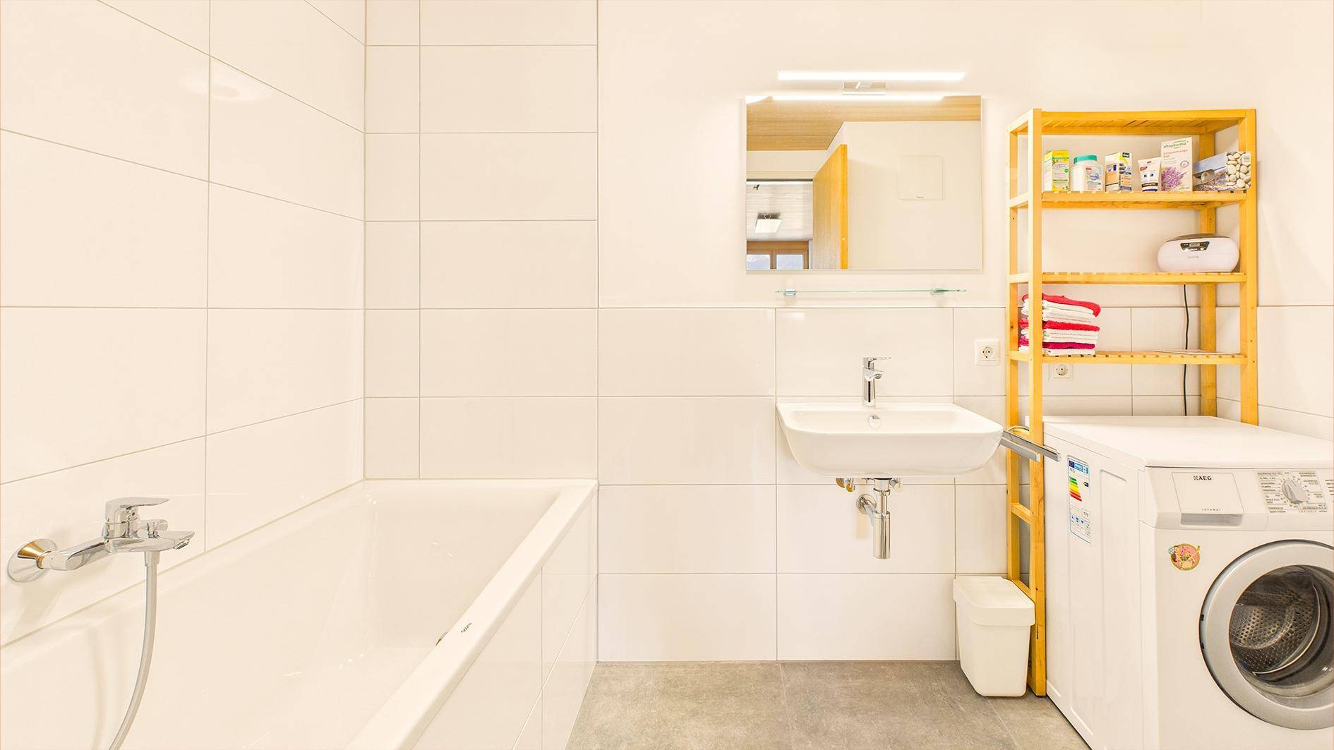 Badezimmer mit Badewanne, Waschbecken, Waschmaschine und weißem Fliesenboden.