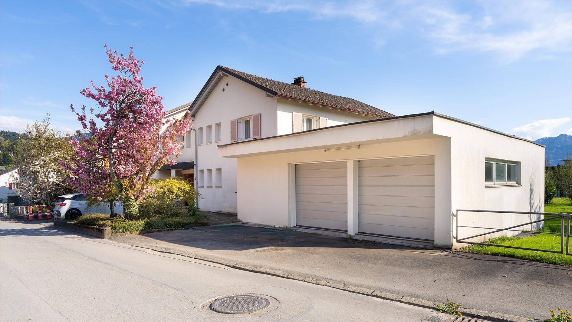 Außenansicht eines weißen Hauses mit Doppelgarage, blühendem Baum und Bergpanorama im Hintergrund.