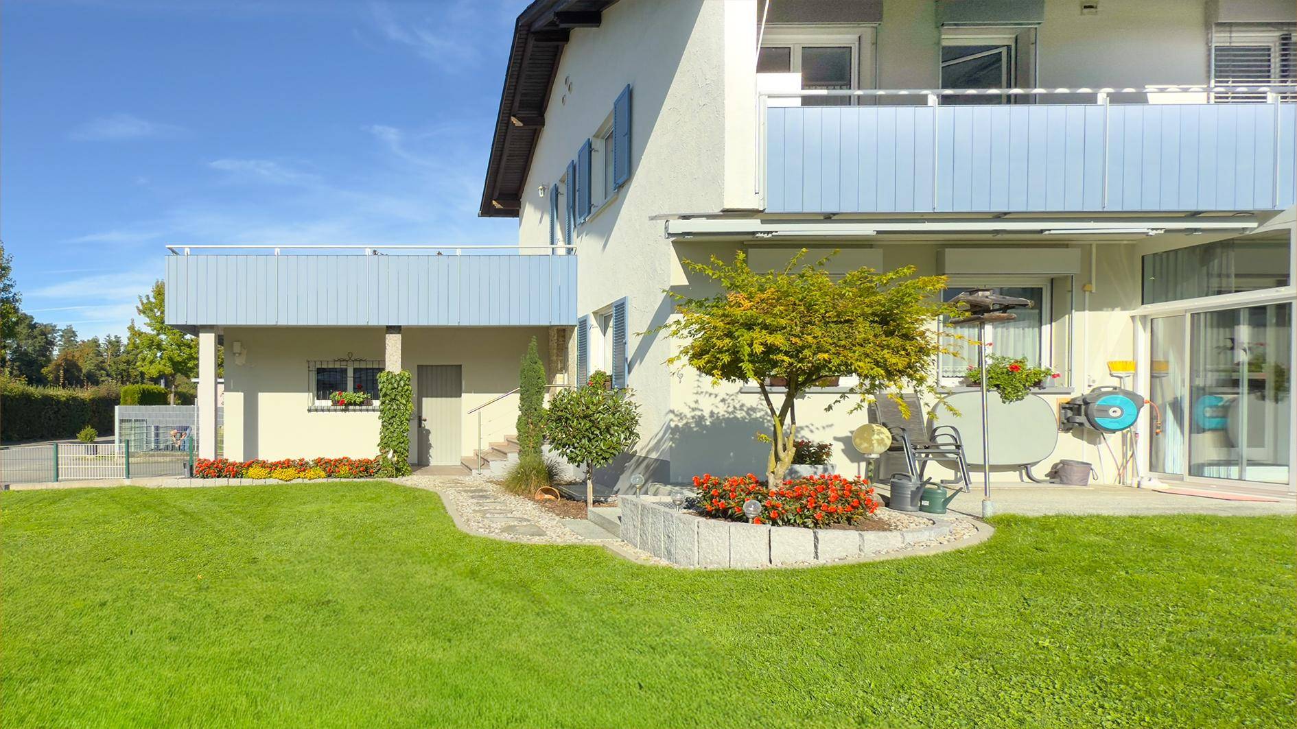 Seitlicher Blick auf das Haus mit gepflegtem Garten, Blumenbeeten und Zugang zur Terrasse.