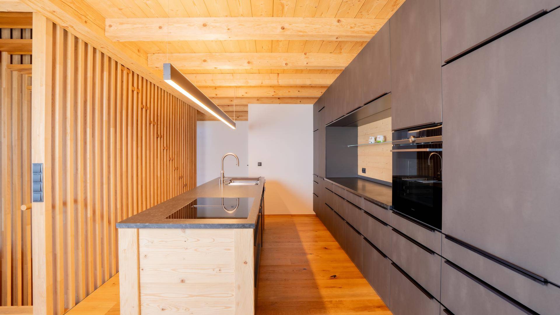Elegante Küche mit dunklen Fronten, Holzinsel und integrierten Geräten unter Holzbalkendecke.