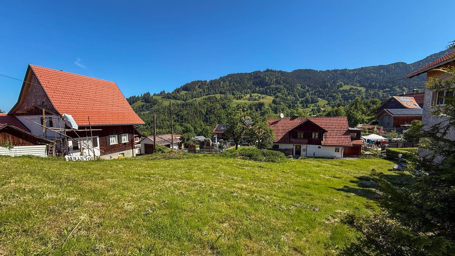 Grünes Baugrundstück mit Blick auf umliegende Häuser und bewaldete Berge im Hintergrund.
