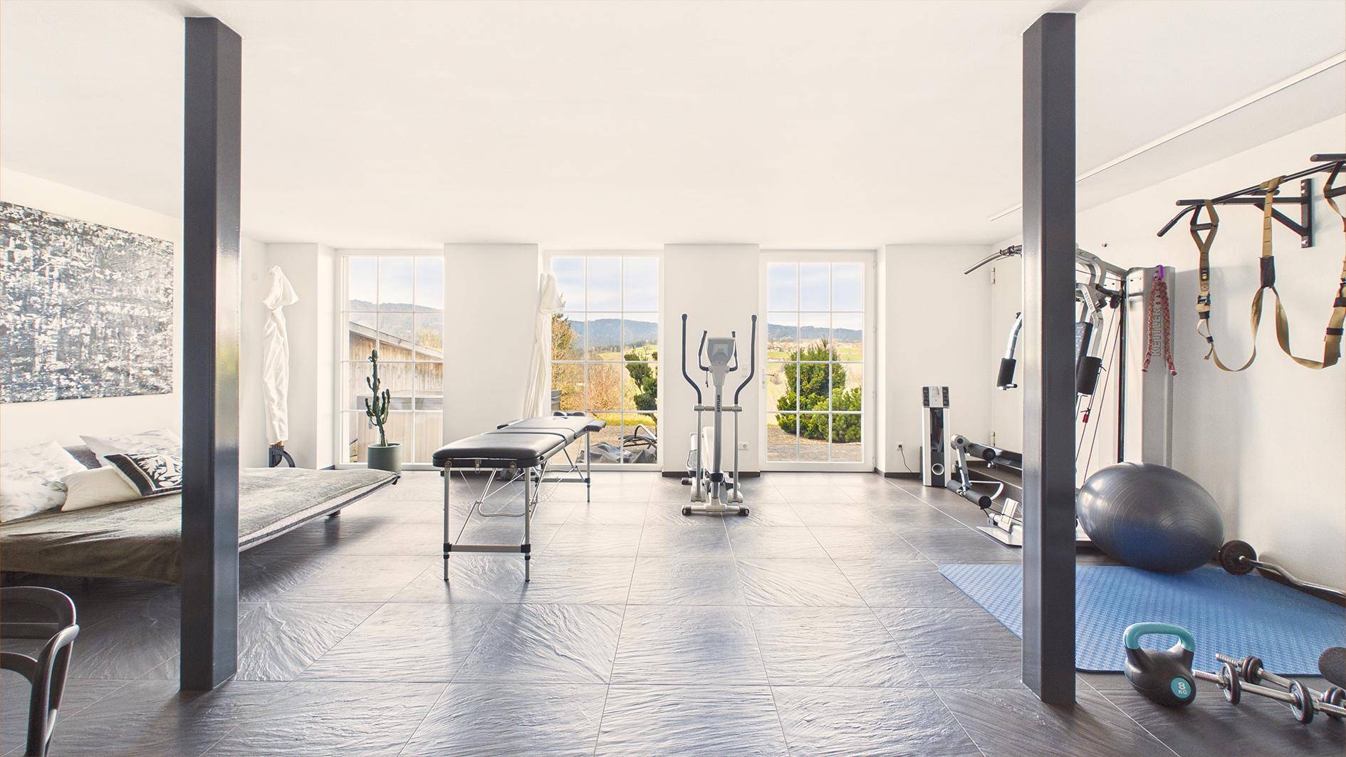 Geräumiger Fitnessraum mit großen Fenstern, dunklem Fliesenboden und Blick ins Grüne.