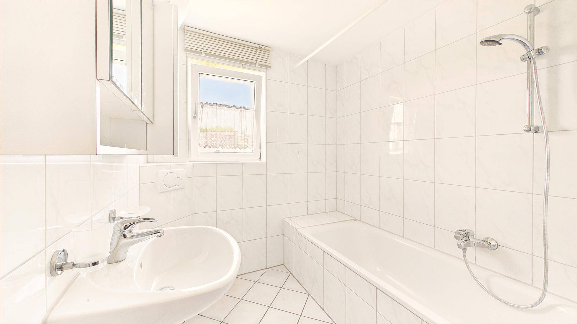 Weiß gefliestes Badezimmer mit Badewanne, Duschvorrichtung und Waschbecken mit Fenster.