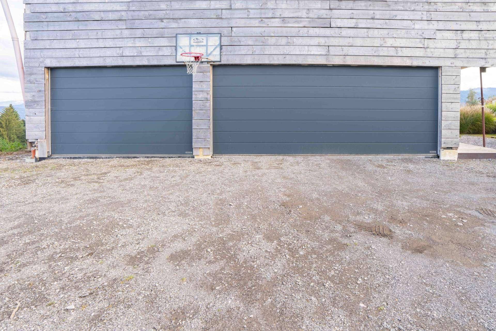 Zwei Garagentore mit Holzfassade und einem Basketballkorb davor auf Schotterfläche.