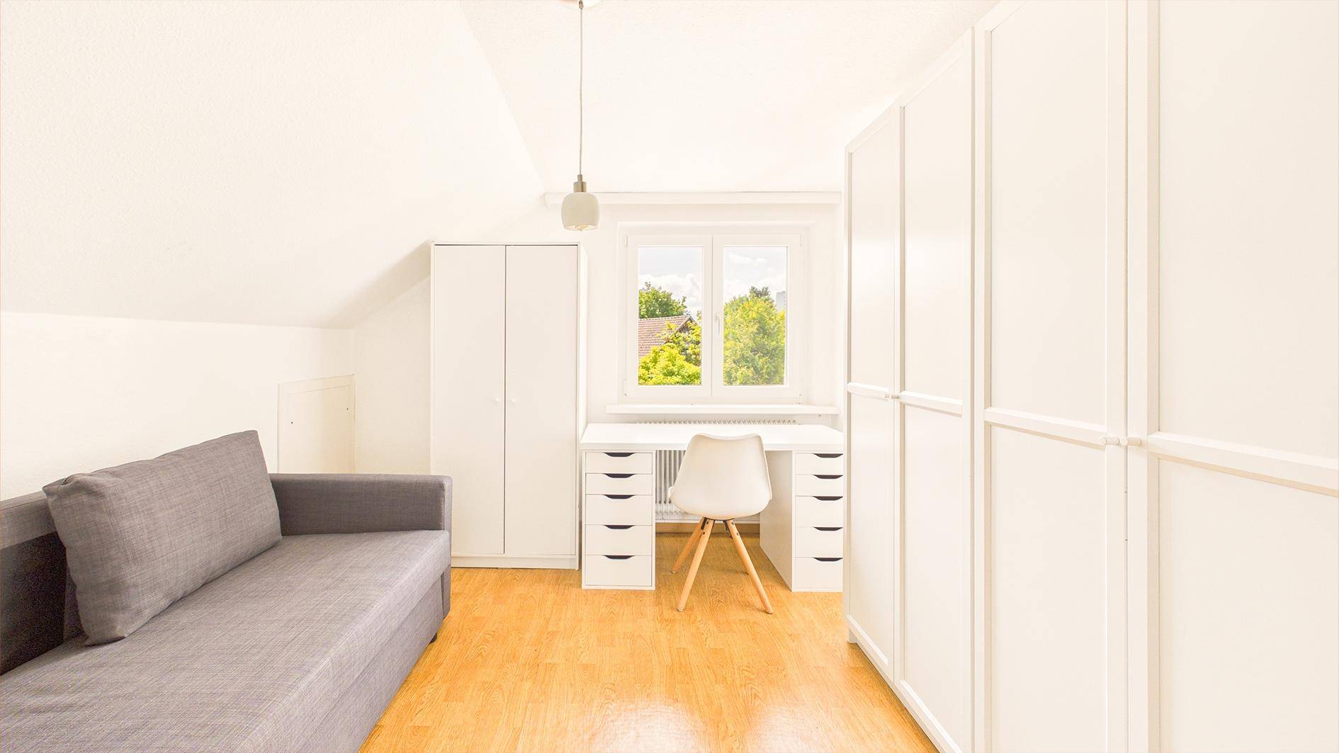 Helles Dachzimmer mit grauem Sofa, weißem Schreibtisch und Einbauschränken, ideal als Schlaf- oder Arbeitszimmer.