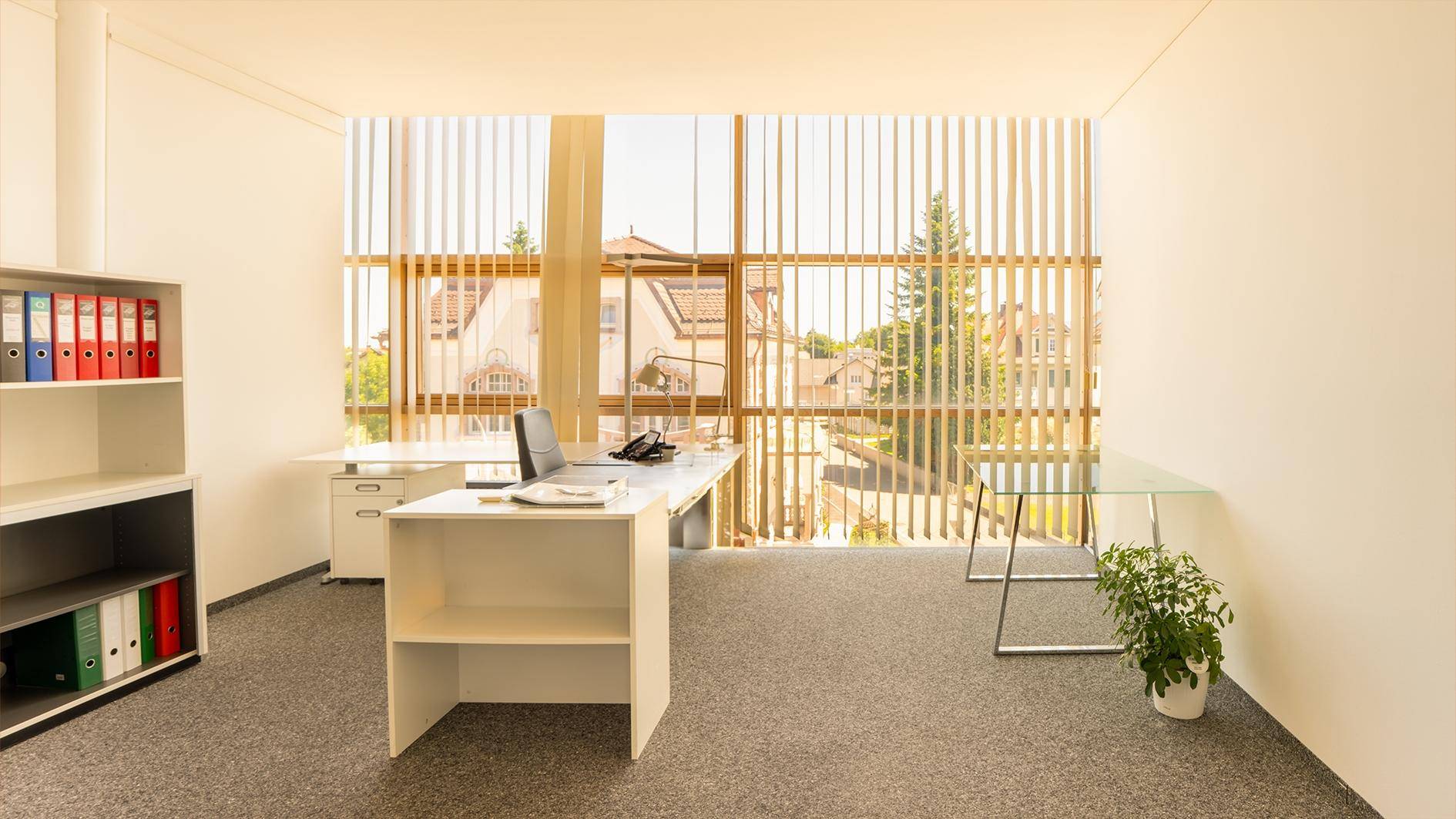 Geräumiges Büro mit großen Fenstern und vertikalen Jalousien, ausgestattet mit Schreibtischen und Regalen.