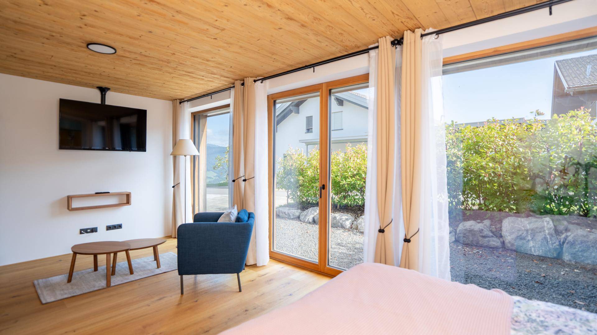 Modernes Schlafzimmer mit Holzboden, Wandfernseher und Blick ins Grüne durch große Fenster.