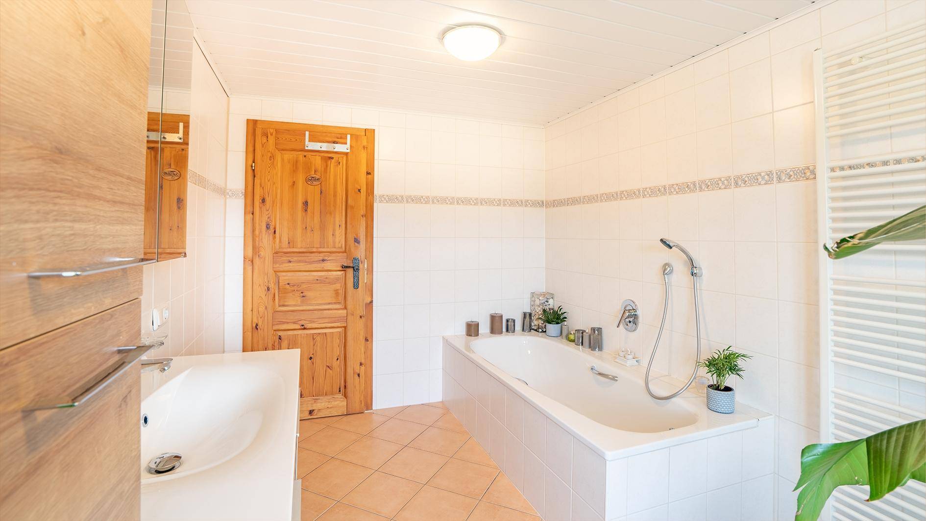 Helles Badezimmer mit Badewanne, Fliesenboden und Holzdetails an der Tür und dem Waschtisch.