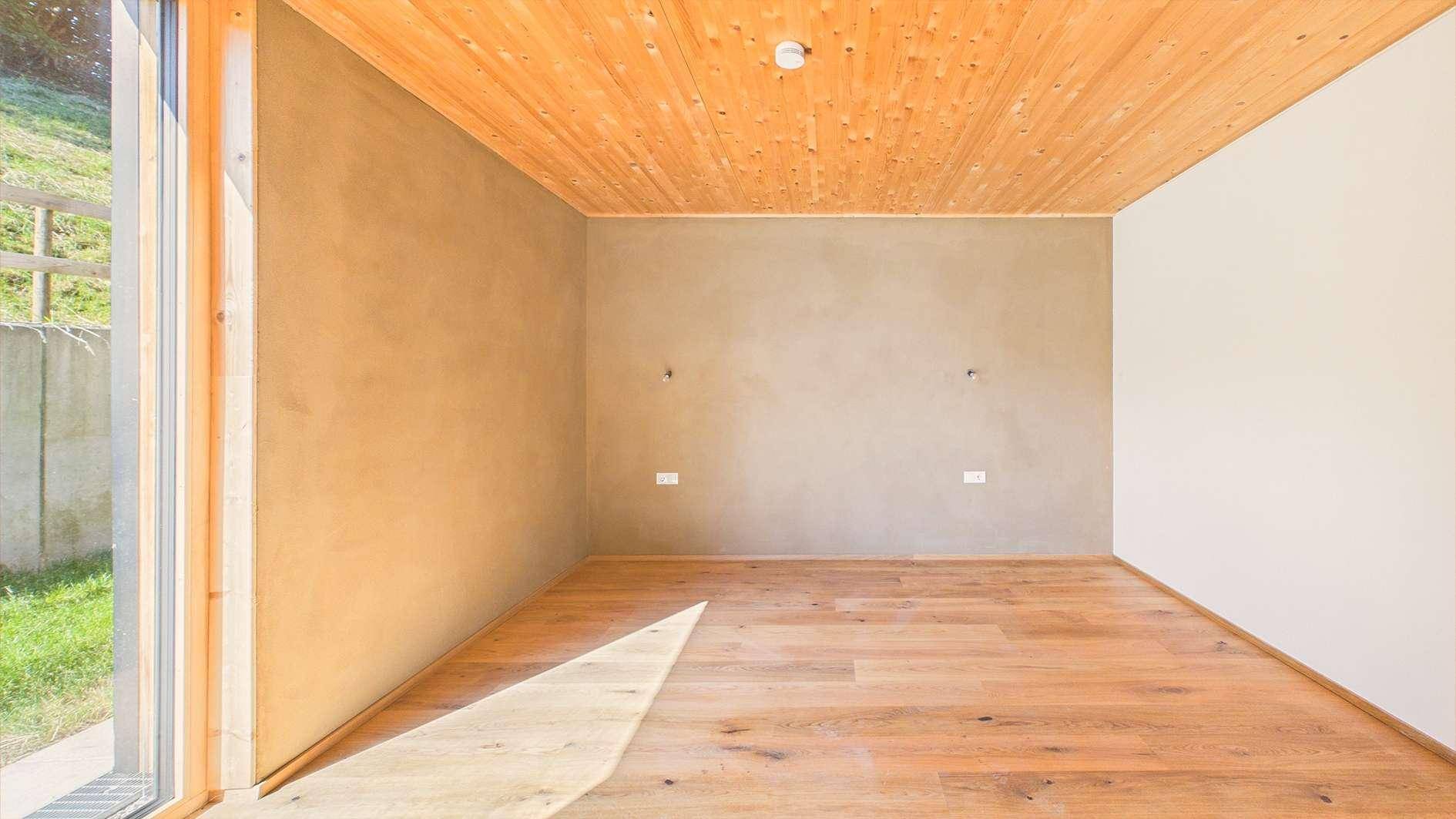Sonniger Raum mit Holzboden und Holzlamellendecke, große Fensterfronten lassen viel Licht herein und bieten Aussicht.